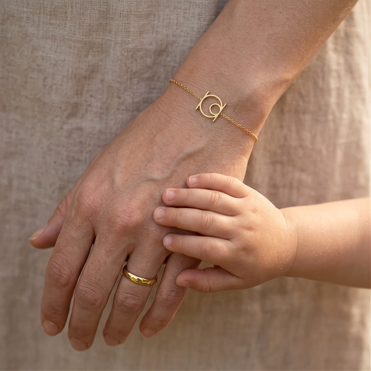 A close-up lifestyle shot of a delicate 9k yellow gold chain bracelet with a Danibydsgn custom single symbol worn on a woman's wrist, with a child's hand resting gently on her hand. Made to order, custom hand made Danibydsgn bracelet.