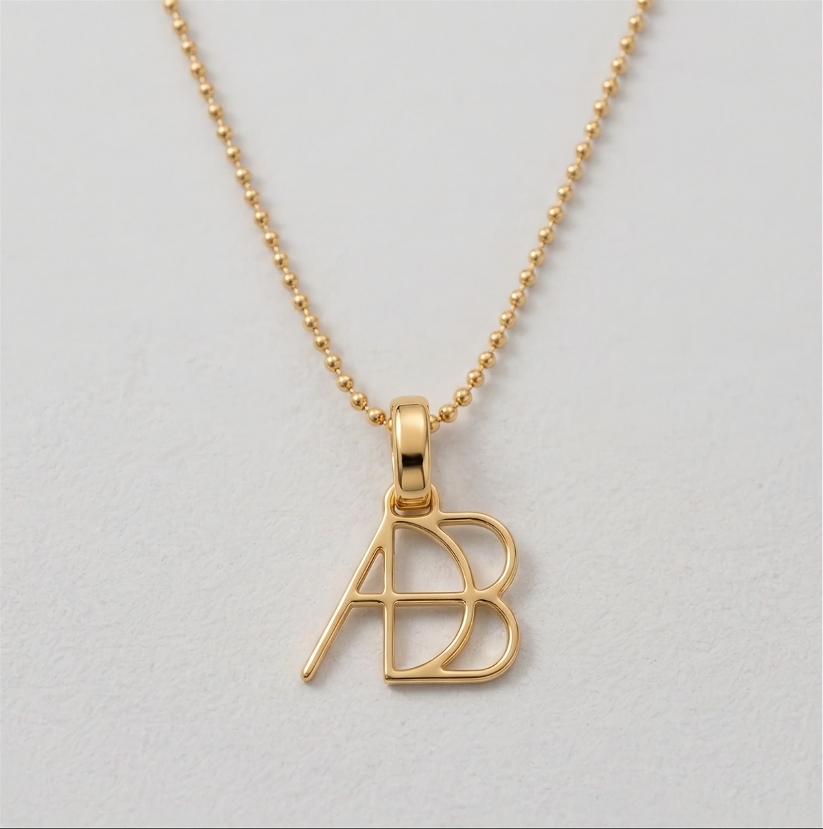 A close up of a 9k gold ball chain necklace and custom Danibydsgn design clip-on charm photographed on a a white textured surface. Meaningful personalised jewelry from Danibydsgn.