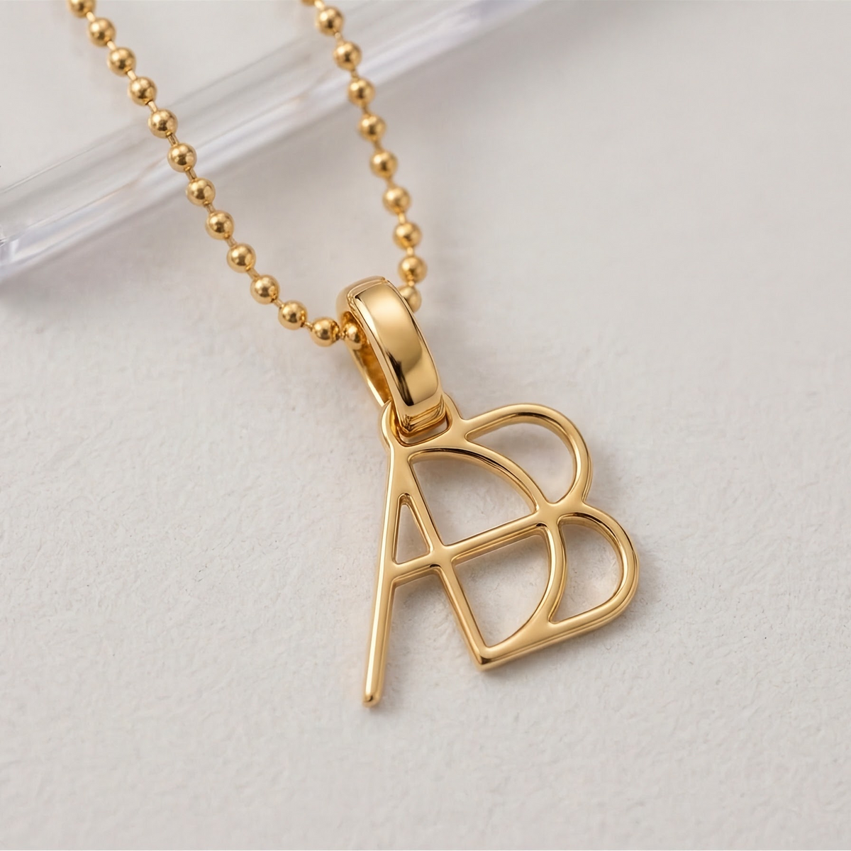 A gold ball-chain necklace with a Danibydsgn design clip on charm in 9k gold photographed on a light neutral textured surface. Elegant meaningful jewelry from Danibydsgn.