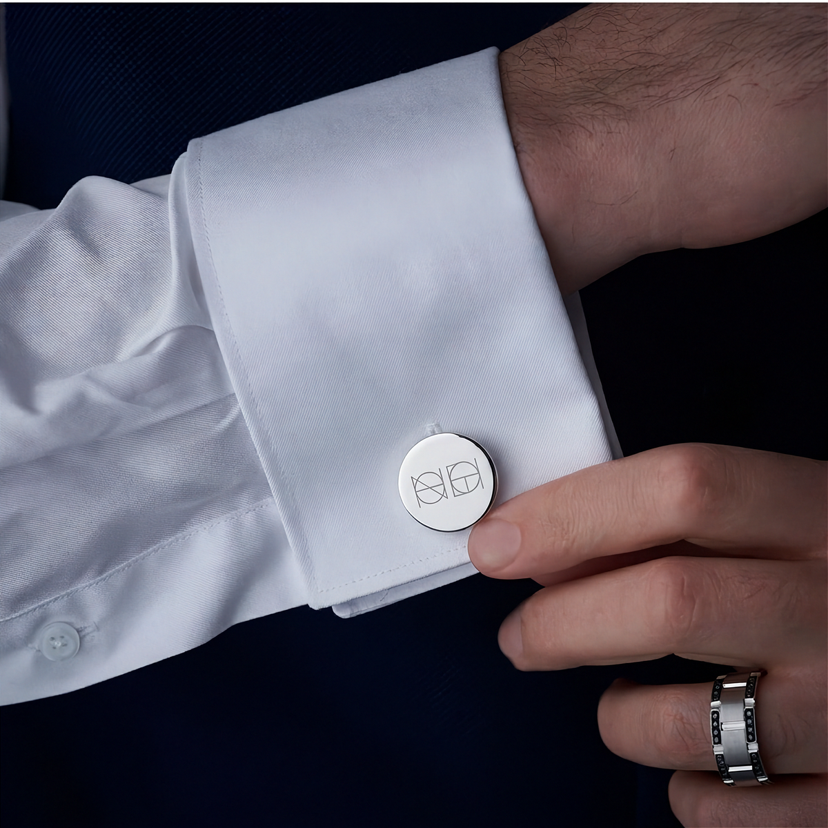 A close-up of a white dress shirt cuff with a round silver Danibydsgn design custom engraved cufflink worn on a mans wrist with a hand adjusting the cuff.