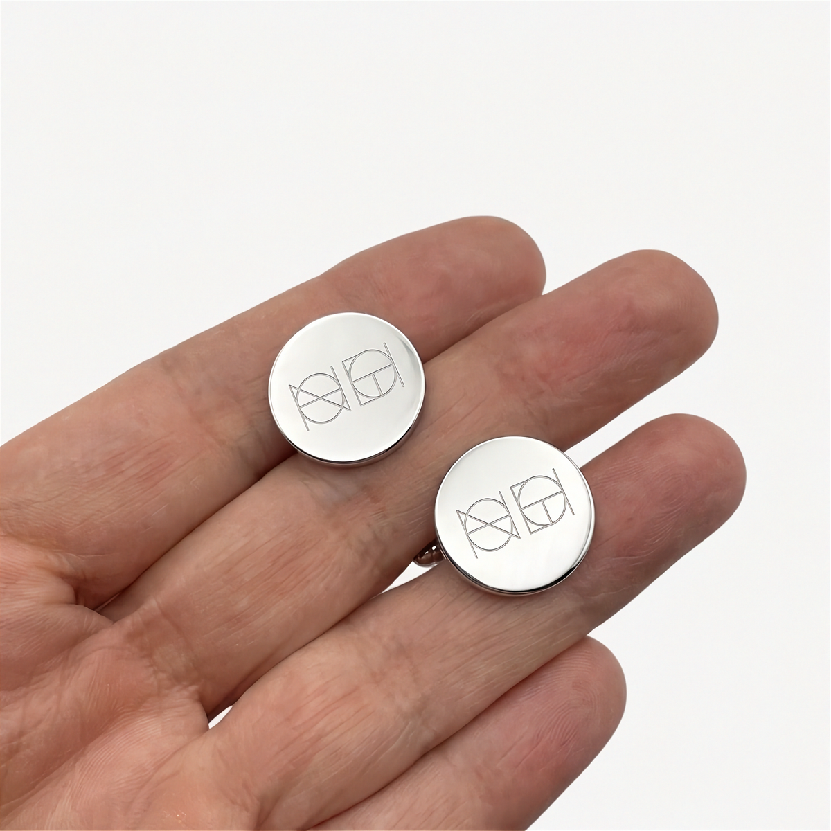 Two round 925 sterling silver cufflinks with a Danibydsgn custom geometric symbol engraved in the center, held between a man's fingers against a plain white background. Two symbol engraved custom Danibydsgn cufflinks.