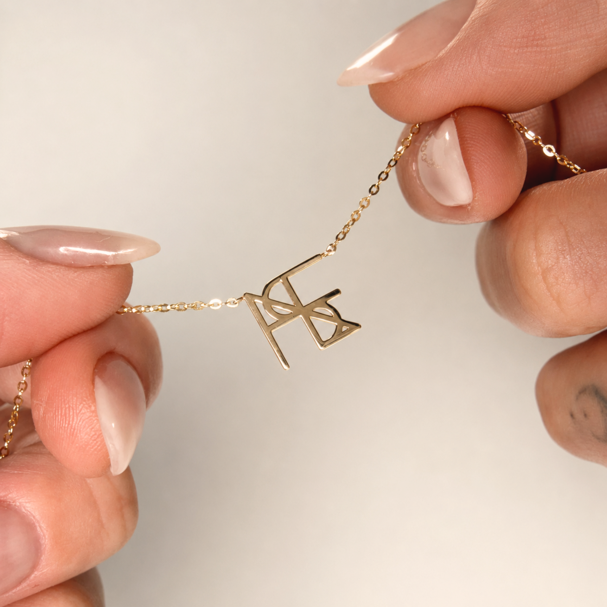 A detailed macro product shot of a model's fingers holding up the delicate chain of a Danibydsgn single symbol pendant necklace in 9k solid gold. The close-up view highlights a personalized uppercase geometric initial charm handcrafted from a custom line art design, pulled taut against a clean, neutral cream backdrop.