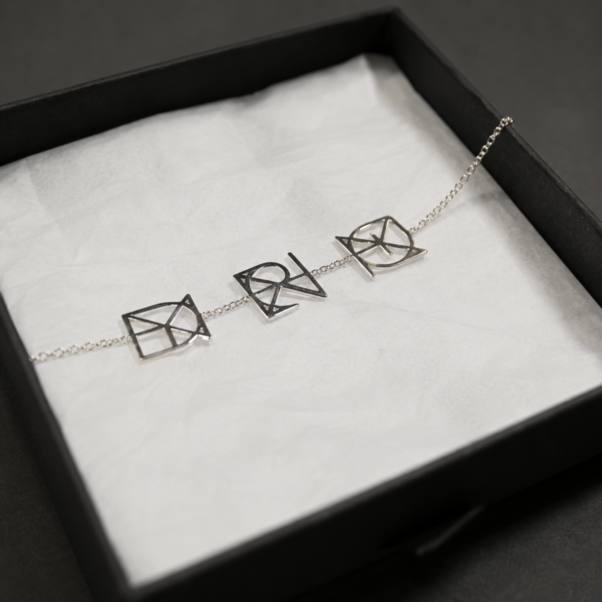 A custom Danibydsgn three symbol pendant bracelet in 925 sterling silver beautifully presented flat inside a black gift box. This made to order modern heirloom charm bracelet features three personalized uppercase geometric charms handcrafted from a customer's meaningful names, words, or dates.
