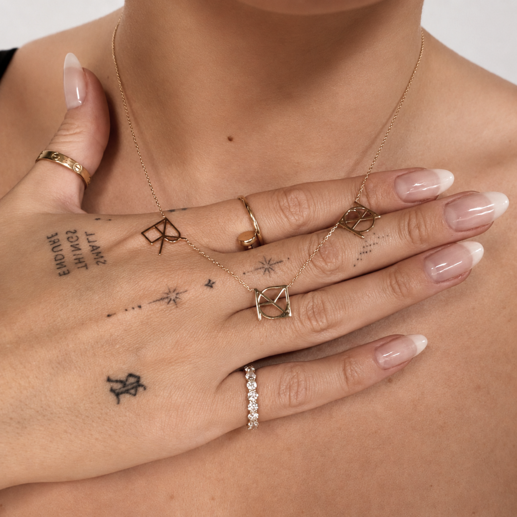 A close-up lifestyle shot of a model wearing a Danibydsgn three symbol pendant necklace in 9k solid gold. This made to order modern heirloom necklace features three personalized uppercase geometric charms handcrafted from a customer's meaningful names, words, or dates on a delicate yellow gold chain, styled gracefully with fine rings and hand tattoos.