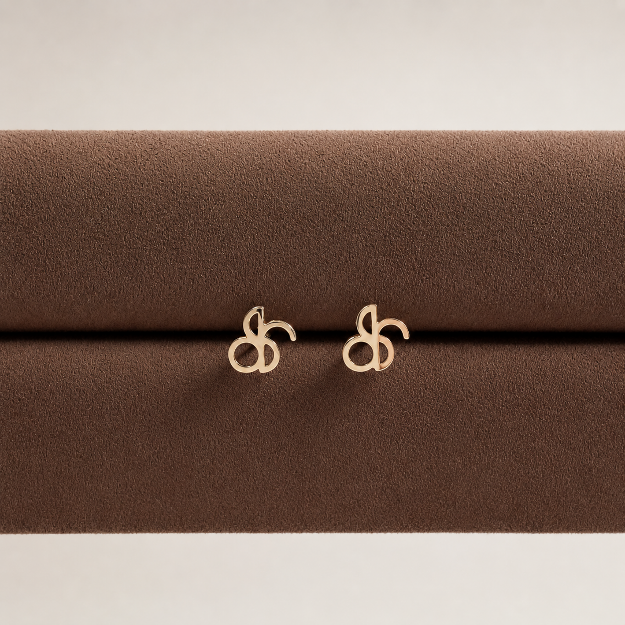 Pair of Danibydsgn lowercase custom fineline design symbol earrings in 9k solid gold. These unique minimalist yellow gold stud earrings feature an abstract lower case design and are displayed on a premium brown Italian velvet jewelry stand, showcasing high-quality, dainty fine jewelry craftsmanship.