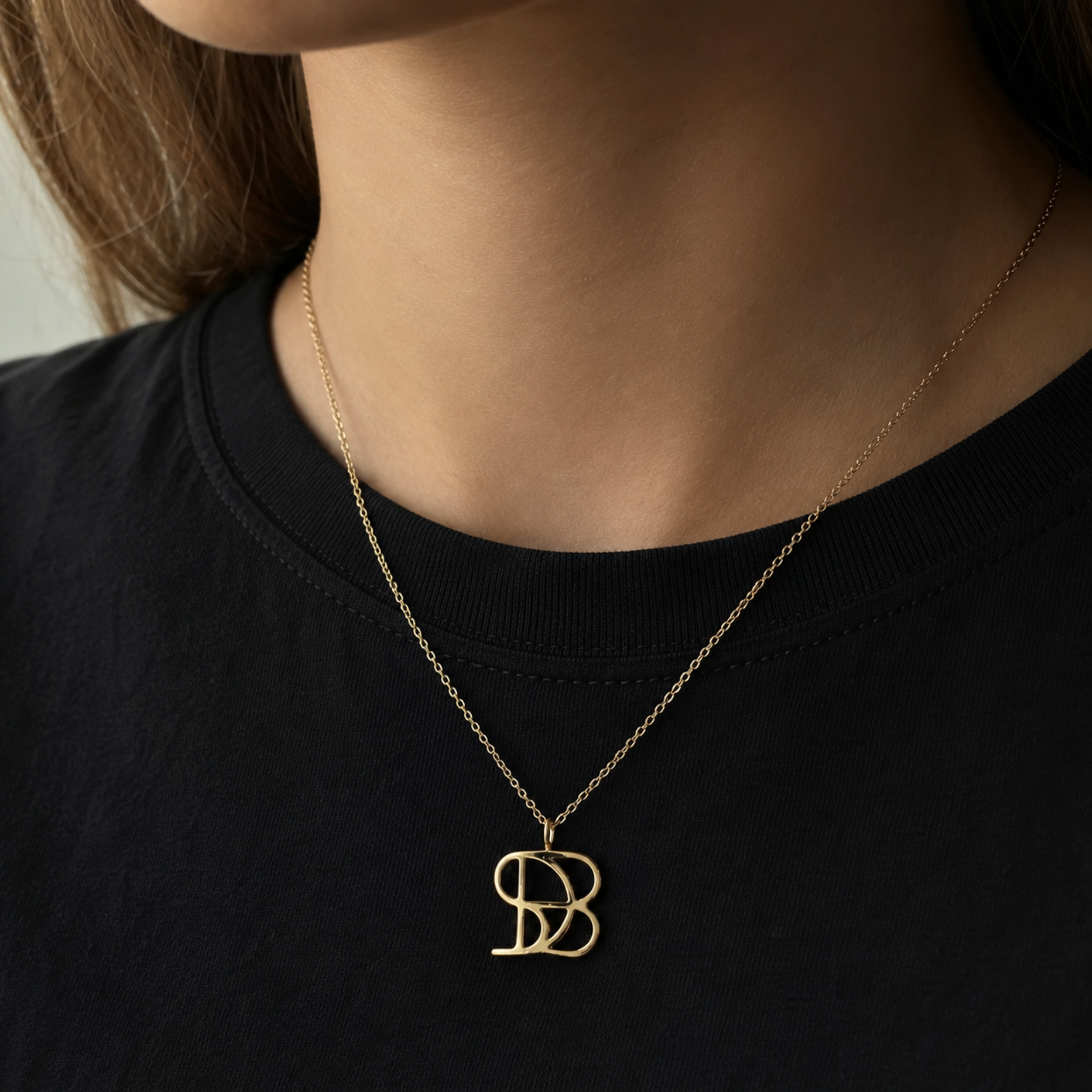 A close-up lifestyle image of a model wearing the Danibydsgn single symbol pendant necklace in 9k solid gold. This personalized architectural necklace features one custom uppercase geometric initial charm handcrafted from a fine-line design, elegantly resting on a delicate yellow gold chain against a classic black t-shirt.