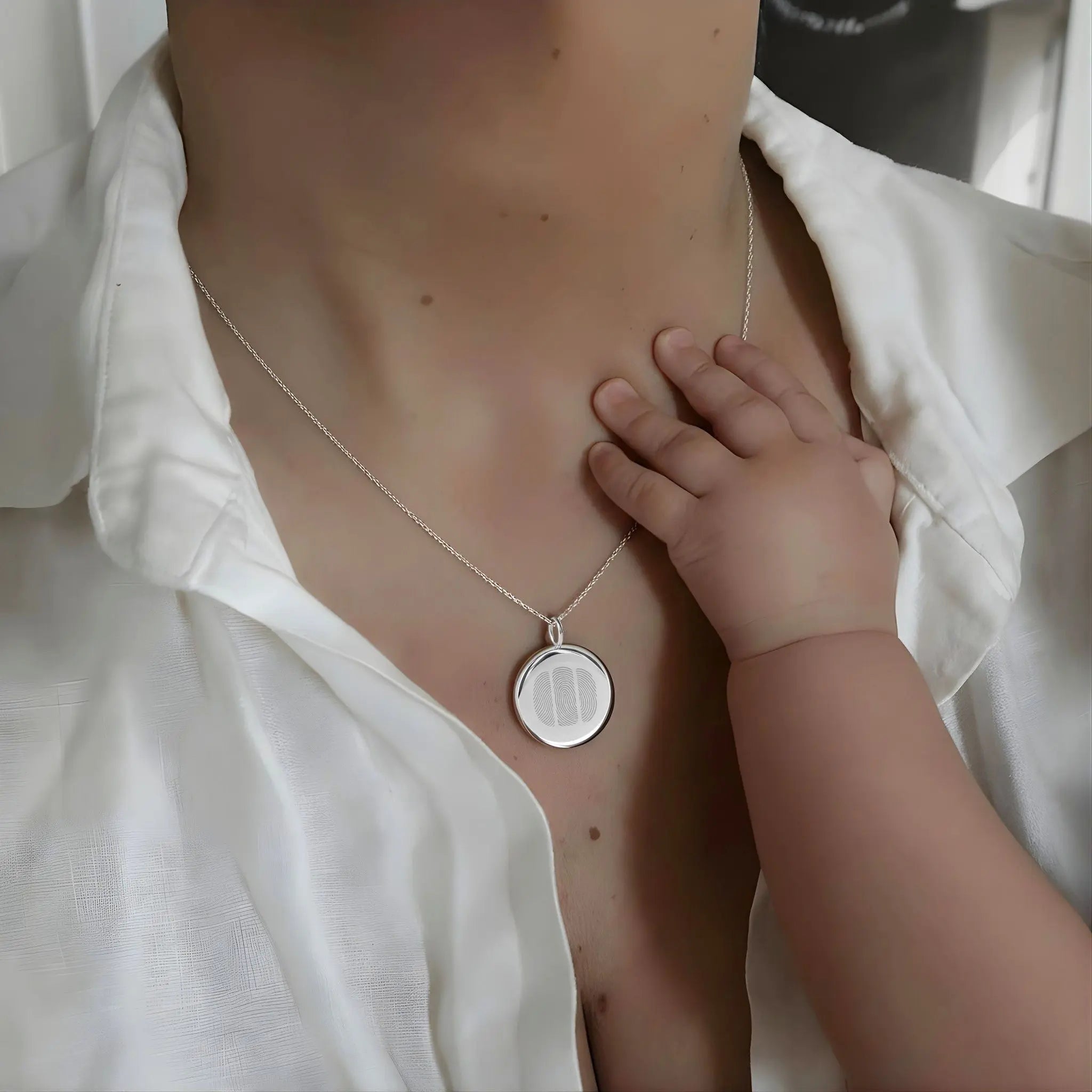 Emotional portrait of a mother wearing the Danibydsgn Three Fingerprint Coin Necklace in 925 Sterling Silver as her baby’s hand rests gently on her chest — symbolising family, love, and everlasting connection captured in timeless craftsmanship.