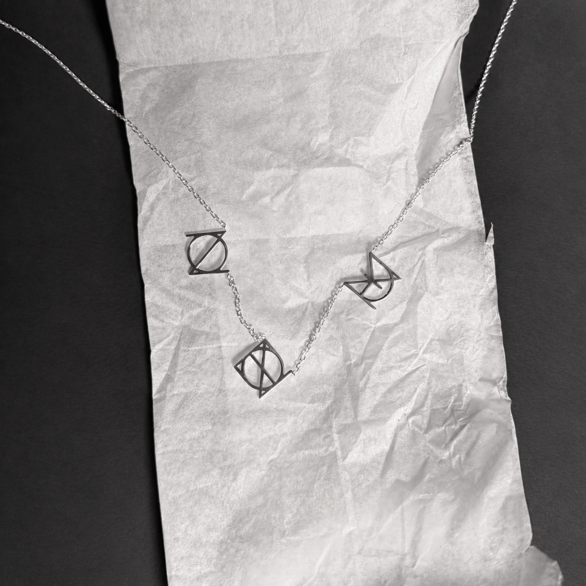 A top-down studio product shot of the Danibydsgn three symbol pendant necklace in 925 sterling silver. This personalized meaningful jewelry piece features three custom uppercase geometric charms on a fine silver chain, laid out neatly on a strip of white crinkled tissue paper against a solid black background.