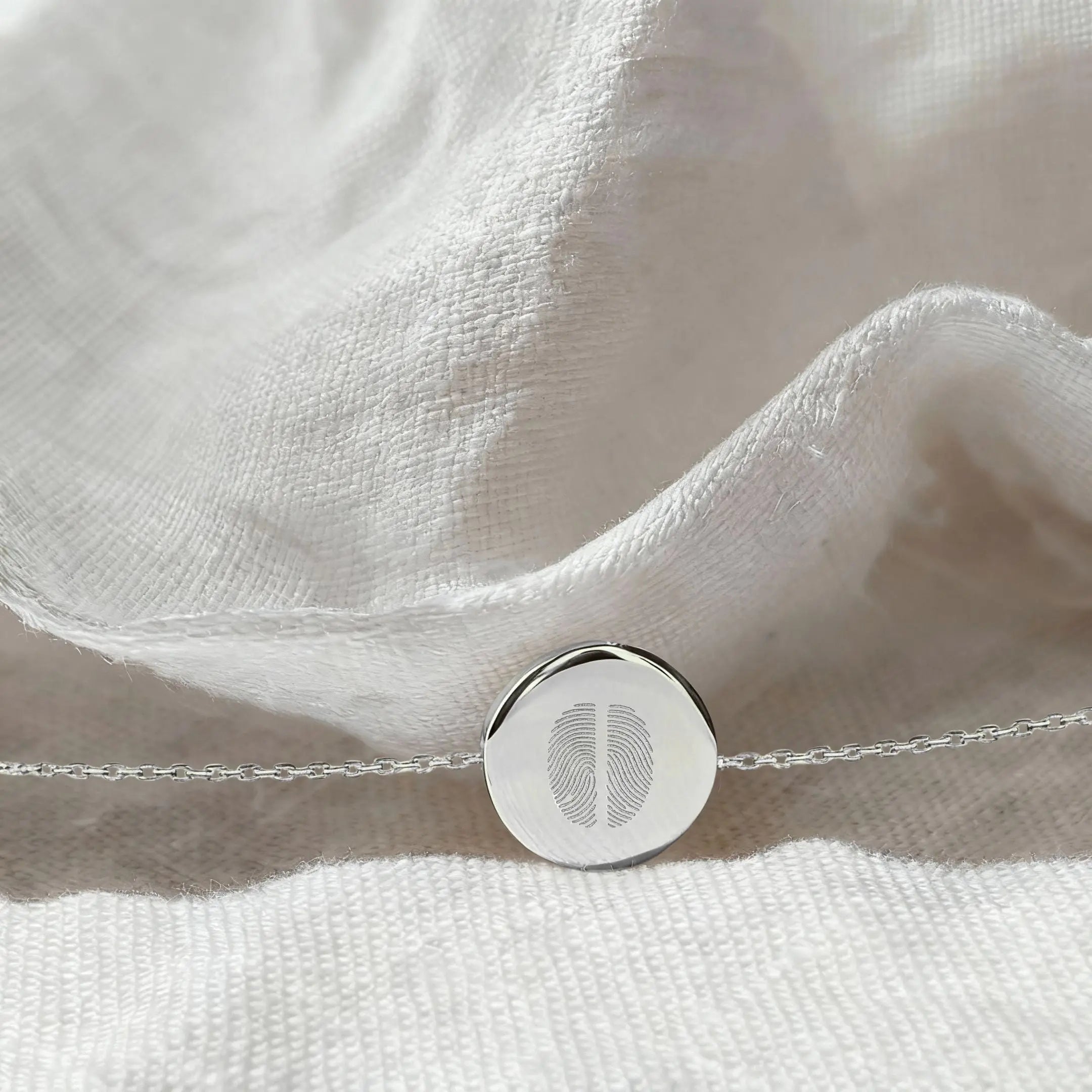 The Danibydsgn Two Fingerprint Coin Bracelet in 925 Sterling Silver resting on softly textured white fabric, highlighting the detailed dual fingerprint engraving and polished round pendant.