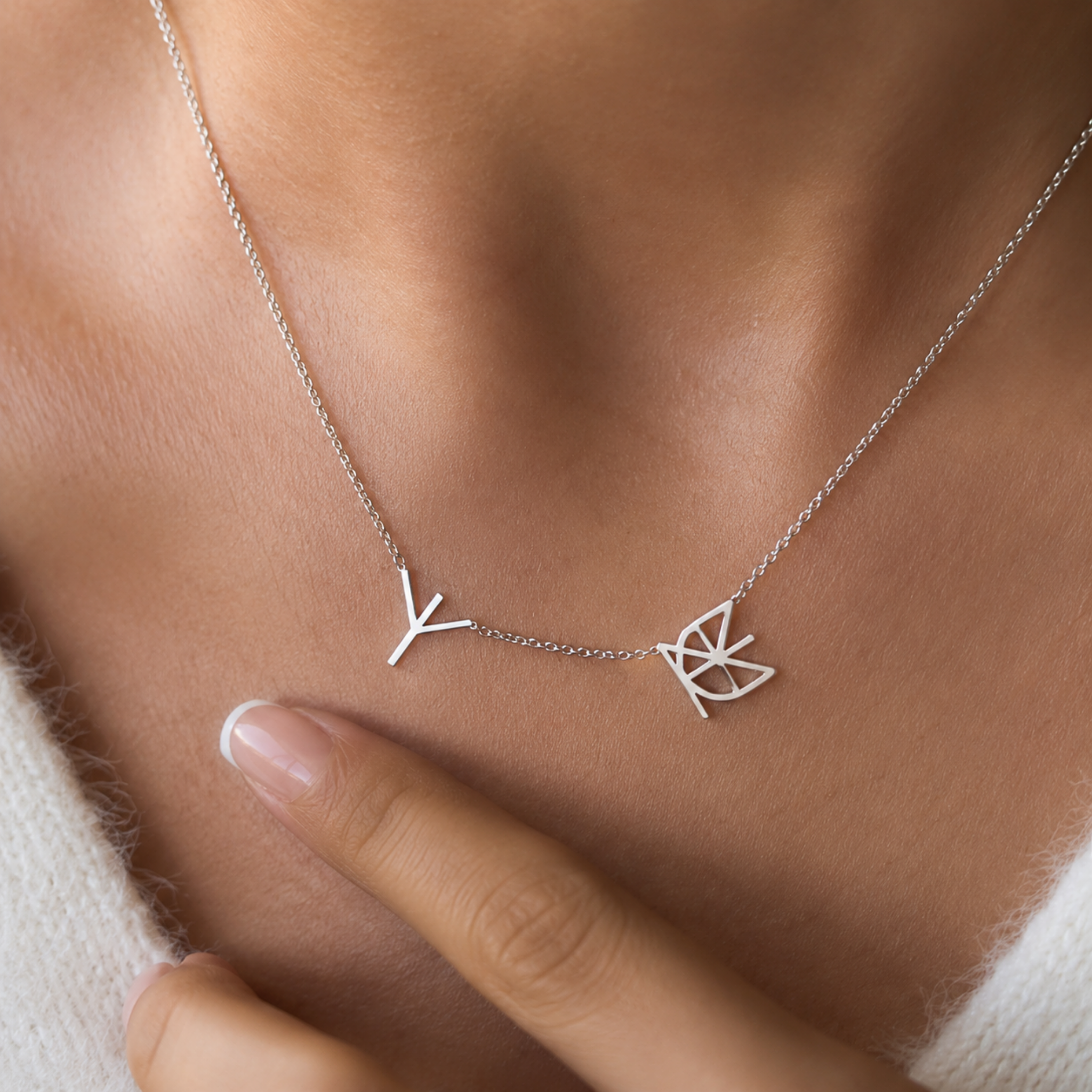A close-up lifestyle image of a model wearing a Danibydsgn two symbol pendant necklace in 925 sterling silver. This minimalist silver necklace features two personalized uppercase geometric initial charms handcrafted from a custom line art design, resting elegantly against the collarbone on a delicate silver chain as a finger touches the textured white knit sweater.