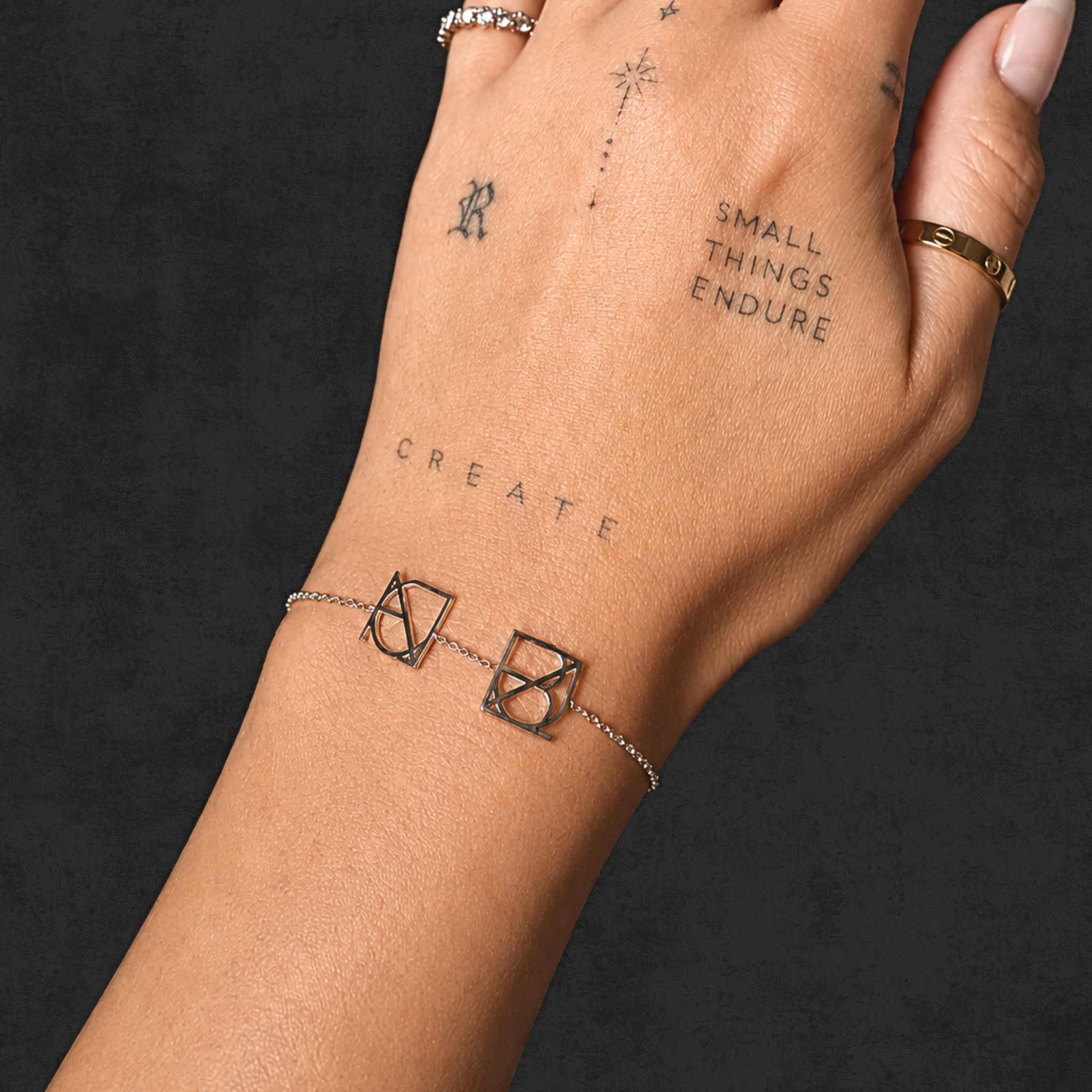 A close-up lifestyle shot of a model's tattooed wrist wearing the Danibydsgn two symbol pendant bracelet in 925 sterling silver. This made to order modern heirloom jewelry features two custom uppercase geometric charms, handcrafted from meaningful names, words, or dates, resting flat on the skin against a dark textured background.