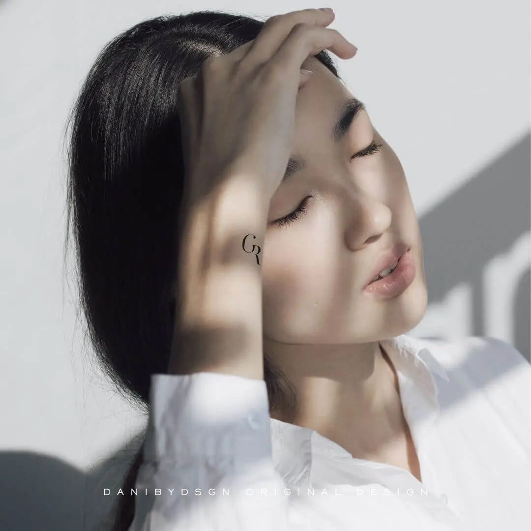 Fine-line initial monogram tattoo design on an arm, featuring minimalist style by Danibydsgn.