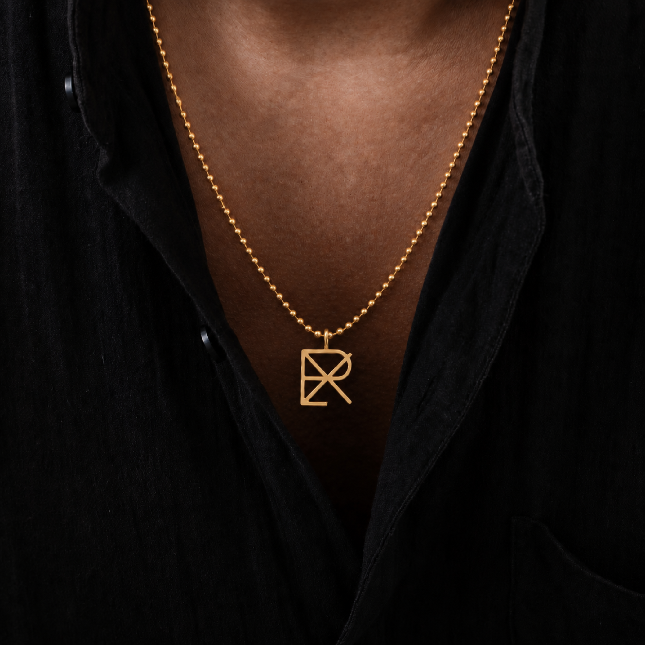 A close-up of a man wearing a 9k gold ball-chain adjustable 20-22 inch chain with a Danibydsgn design cut out pendant, styled with a black button-down shirt.