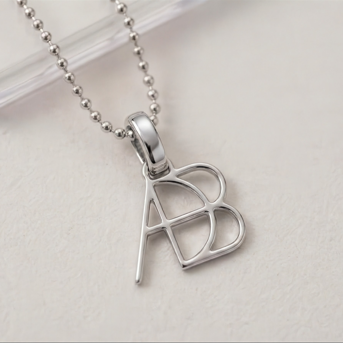 A close up shot of a 925 sterling silver ball chain necklace and custom made to order Danibydsgn design clip-on charm photographed on a light textured surface. Meaningful personalised jewelry from Danibydsgn.