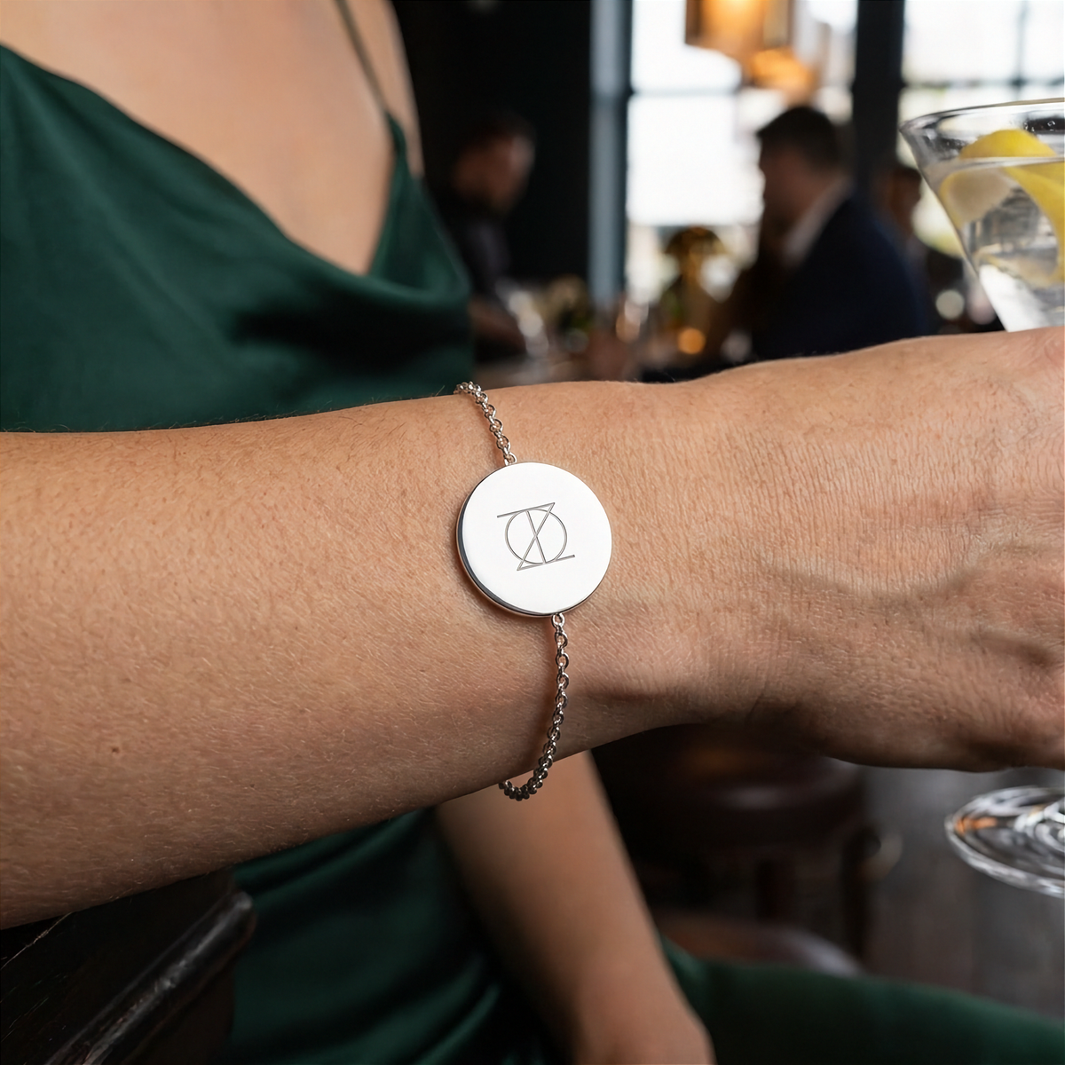 Danibydsgn custom symbol engraved coin bracelet, 925 sterling silver being worn by a lady in an elegant green dress. Single symbol design in upper case letters.