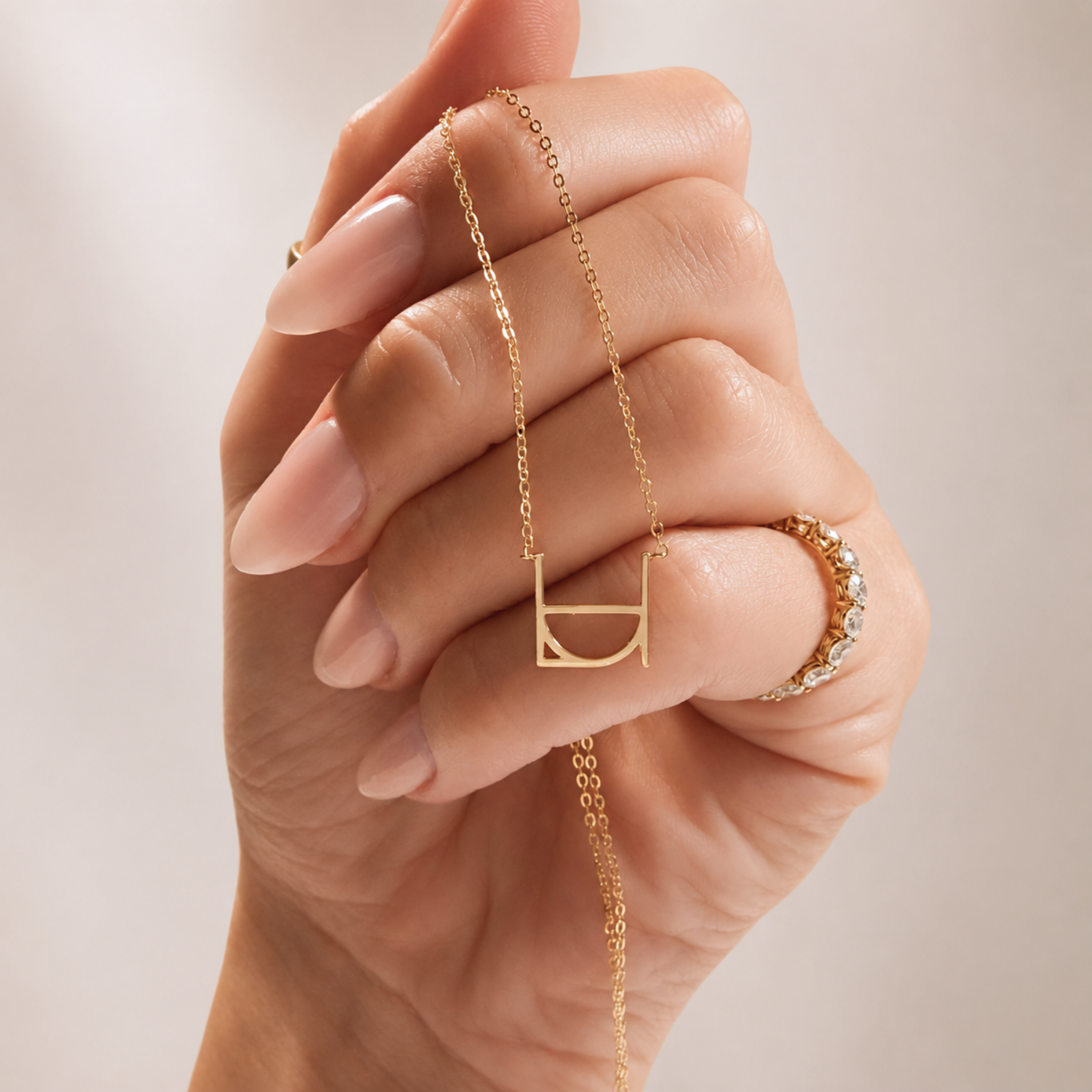 A close-up lifestyle shot of a model's hand holding the Danibydsgn single symbol pendant necklace in 9k solid gold. This dainty 9k gold necklace features one personalized uppercase geometric initial charm handcrafted from a custom line art design, draped elegantly over the fingers to display the delicate yellow gold chain against a soft, neutral background.
