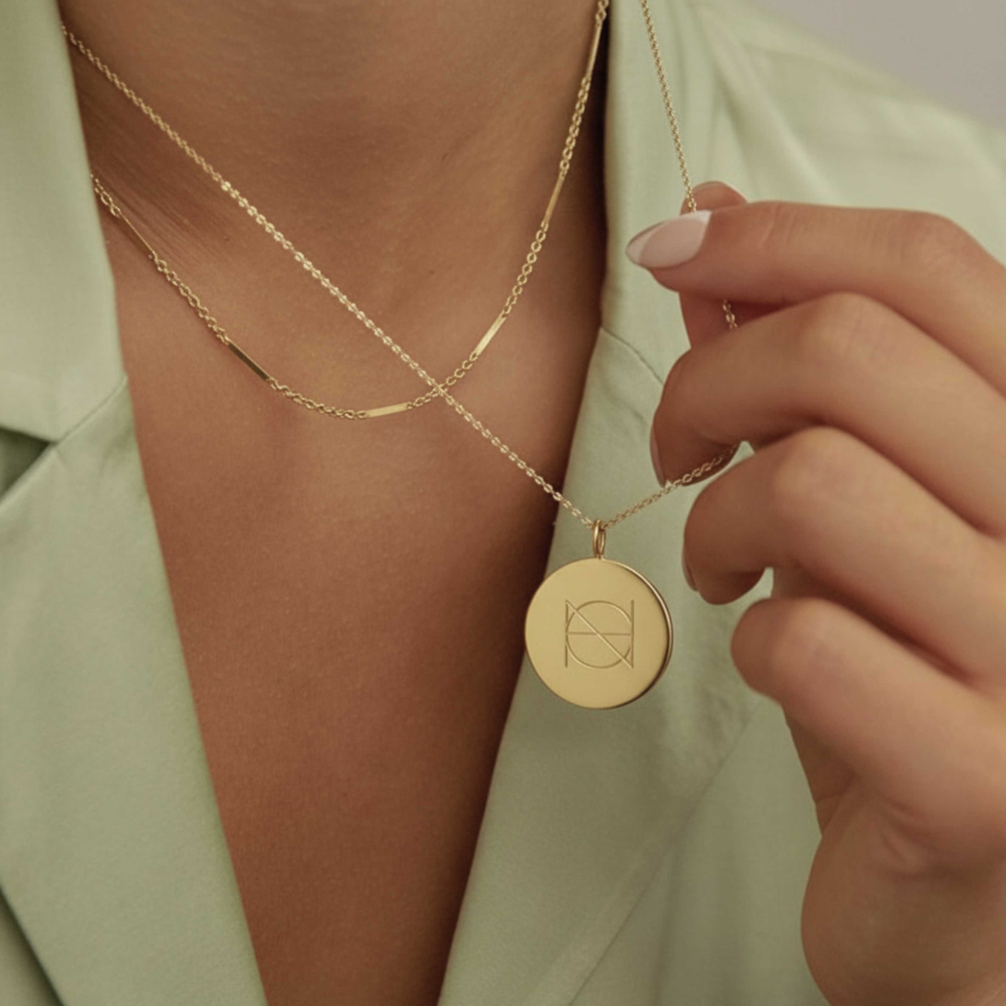 Gold coin pendant necklace  engraved with  a Danibydsgn logo  worn by a lady wearing an elegant sage green silk shirt. 