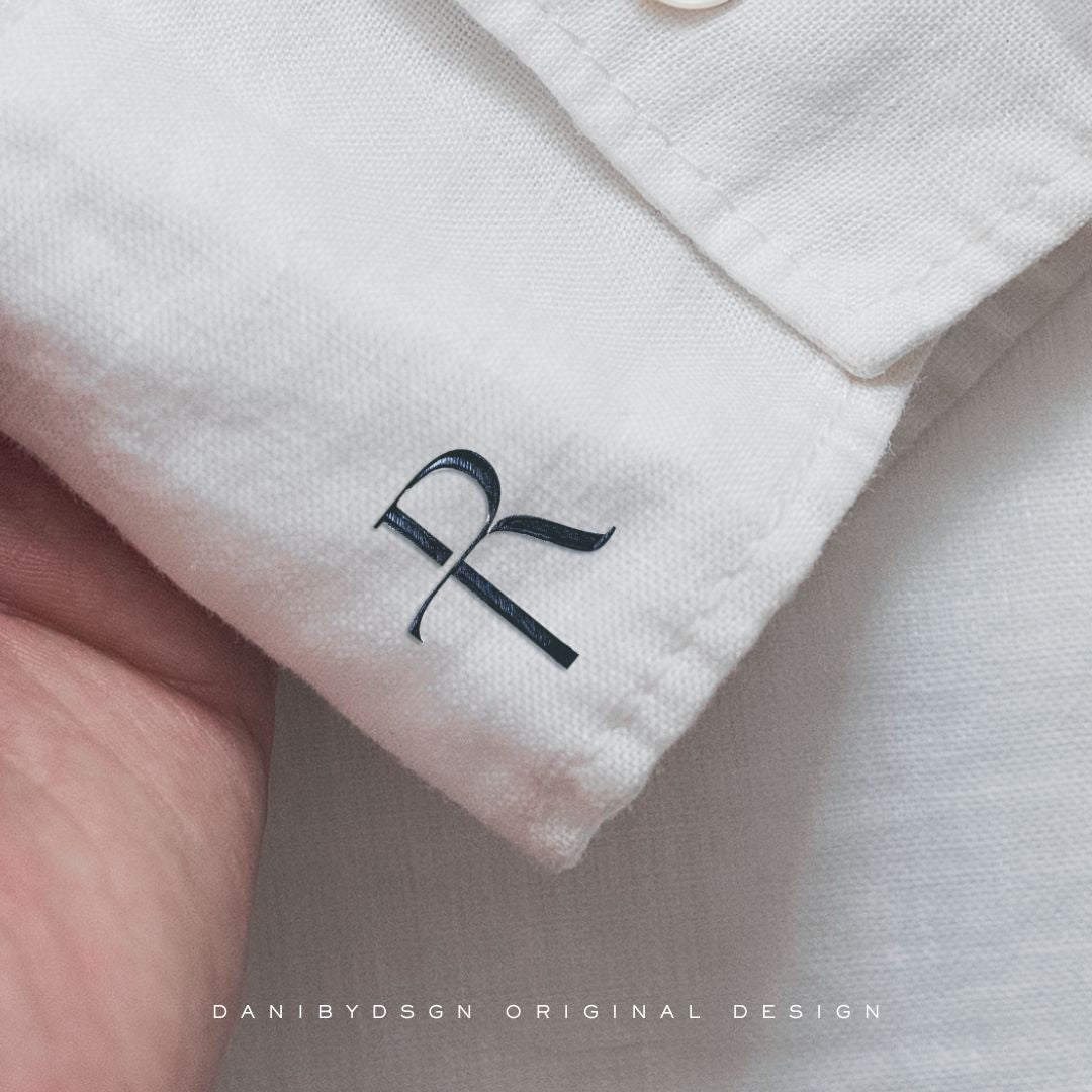 Image of a clothing label with an initial monogram logo representing Business Initial Monogram Design. The monogram is designed by Danibydsgn, showcasing expertise as a business logo designer and graphic designer. This small business logo is a perfect example of professional branding, ideal for clothing branding. The black monogram contrasts against the white fabric, symbolizes sophisticated business branding. This image displays a business initials logo and is ideal as a website brand logo branding.