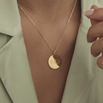 Gold coin pendant necklace  engraved with two Danibydsgn logos  worn by a lady wearing an elegant sage green silk shirt. 