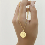 Gold coin pendant necklace engraved with a custom Danibydsgn single symbol design held by a hand against a light background
