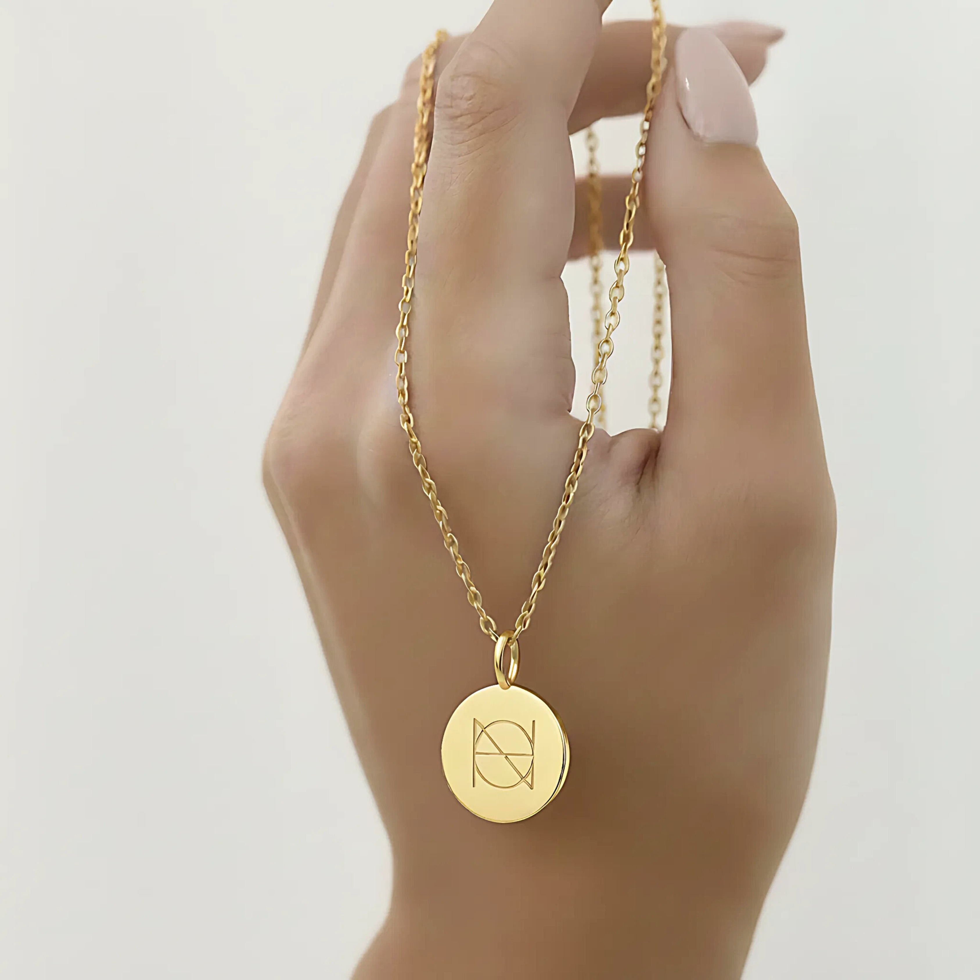 Gold coin pendant necklace engraved with a custom Danibydsgn single symbol design held by a hand against a light background