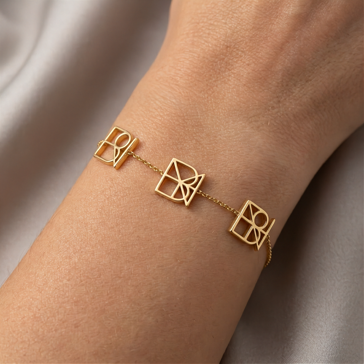 A delicate 9k yellow gold chain bracelet with three Danibydsgn design cut out geometric symbols, shown worn on a person's wrist against a soft beige satin background.  Custom hand made, made to order jewelry.
