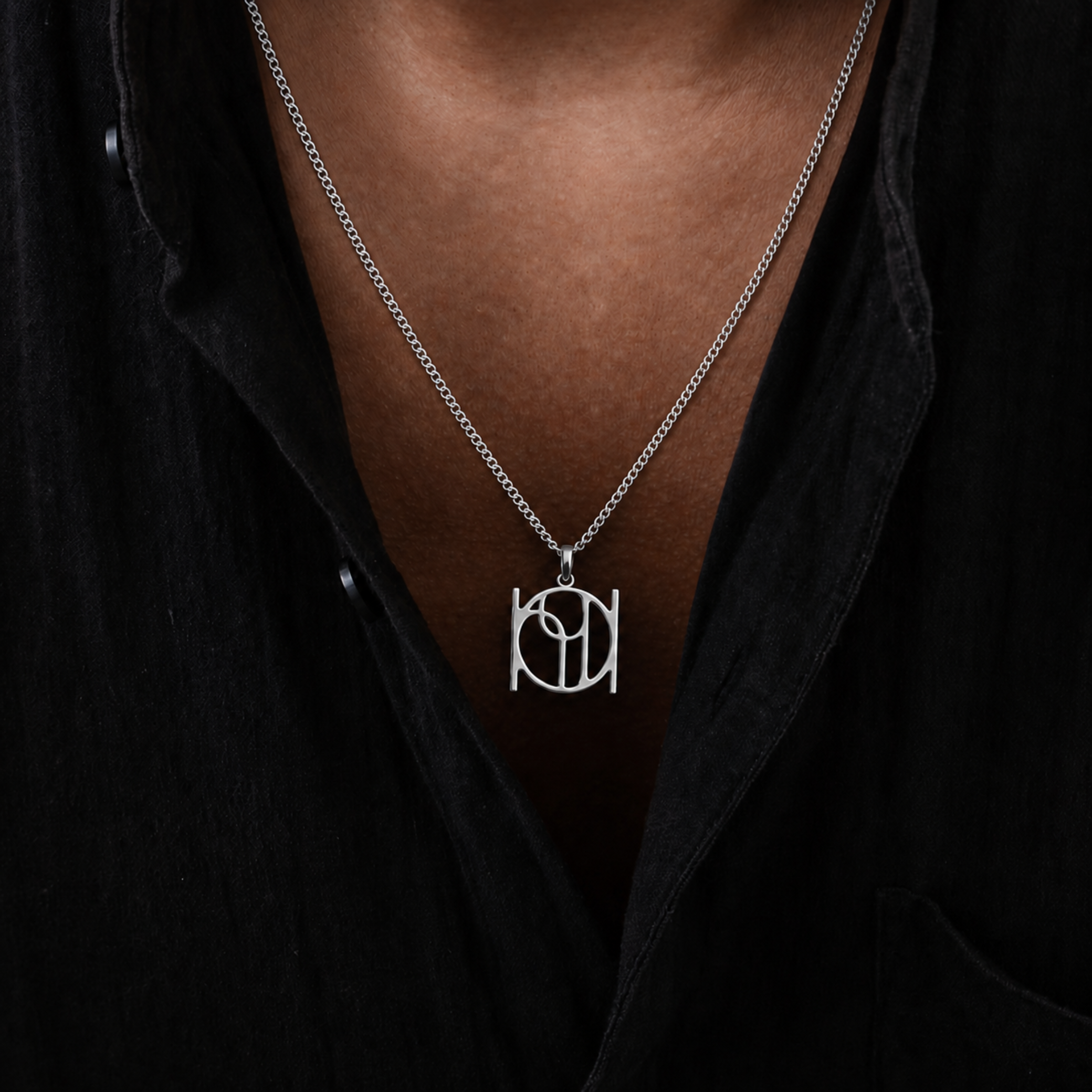 A close-up of a person wearing a silver cable chain necklace with Danibydsgn interlocking letters cut out pendant, styled with a black open-collar shirt.  Luxury jewelry. 