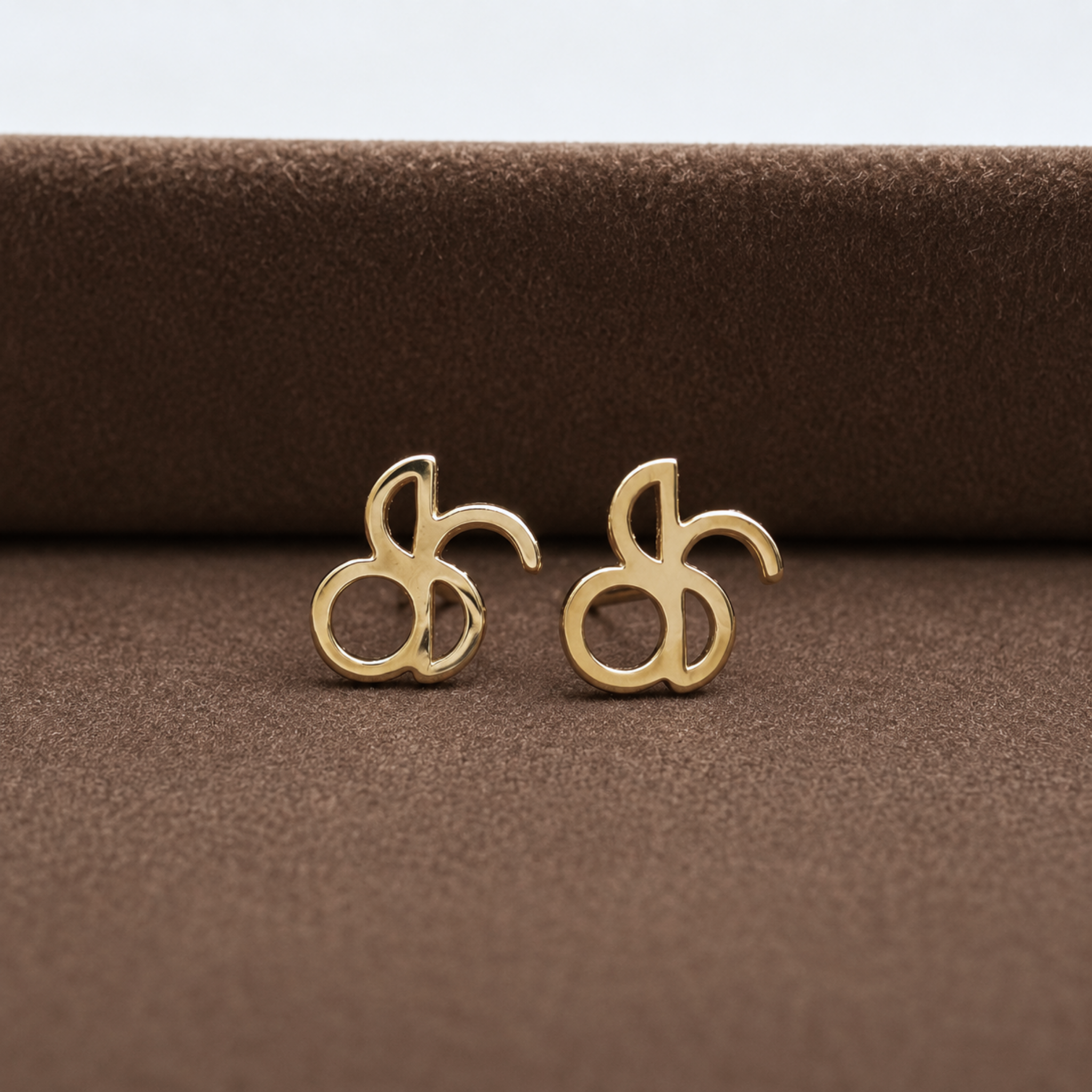 A pair of Danibydsgn custom line art symbol earrings crafted from polished 9k solid yellow gold, featuring an intricate, flowing abstract design. The two minimalist stud earrings are displayed side-by-side on a textured, dark brown plush velvet surface, highlighting the reflective gold metalwork.