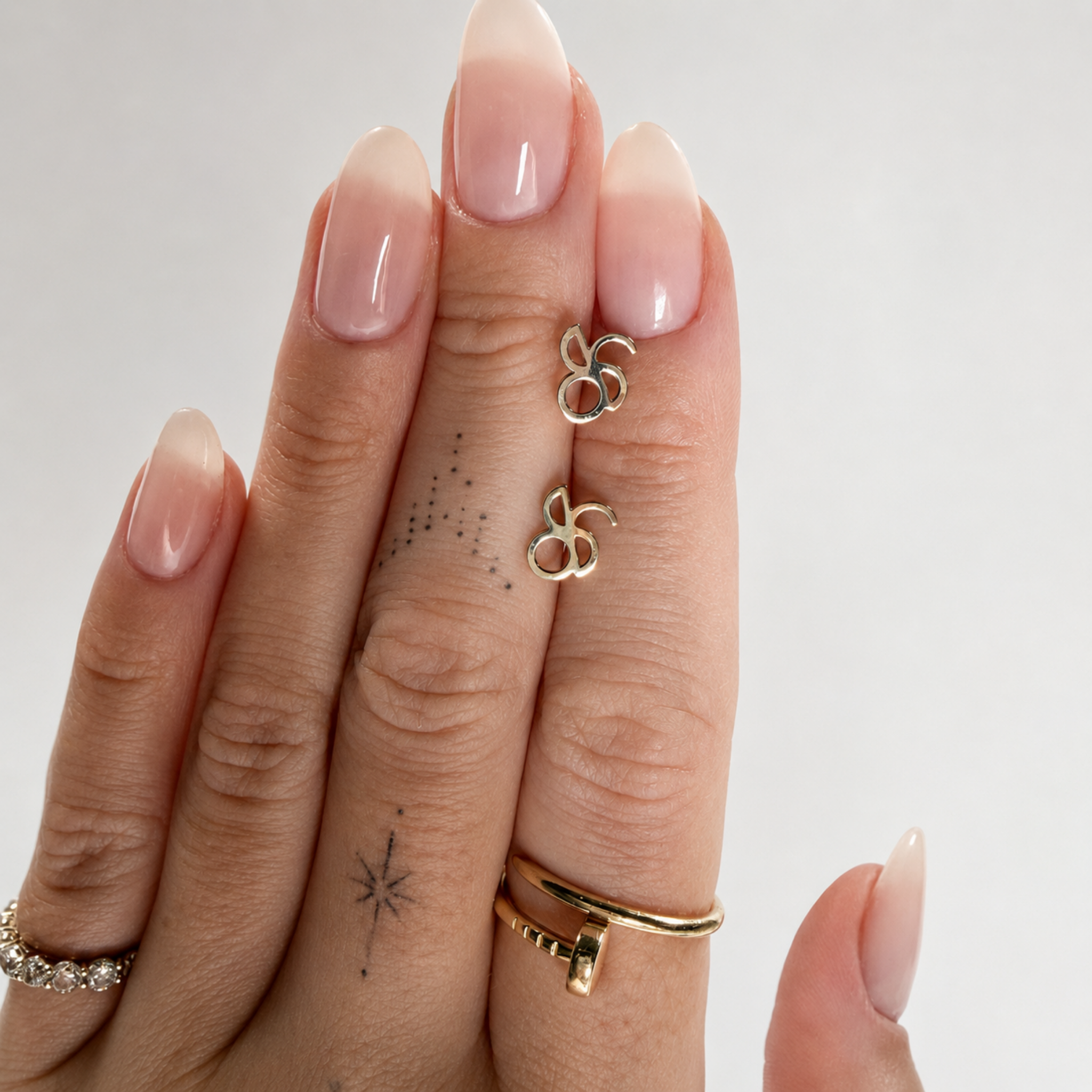 A hand with a clean manicure and delicate finger tattoos holds a pair of Danibydsgn custom line art symbol earrings in 9k gold against a white background, demonstrating the small, dainty scale of the solid yellow gold minimalist studs next to the fingers.