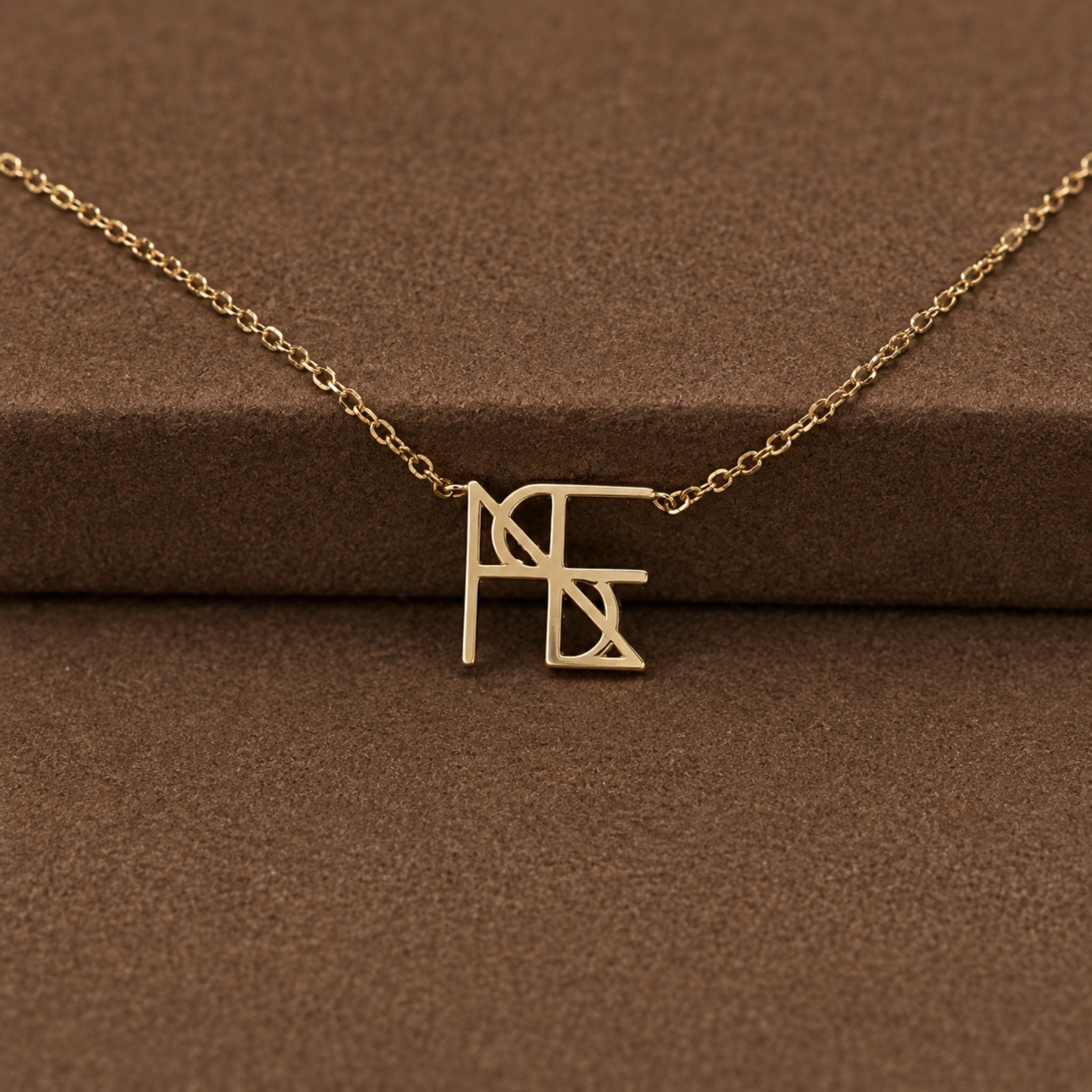 A close-up studio product shot of the Danibydsgn single symbol pendant necklace in 9k solid gold. This dainty yellow gold necklace features one personalized uppercase geometric initial charm handcrafted from a custom line art design, resting elegantly across a textured brown velvet jewelry display surface.