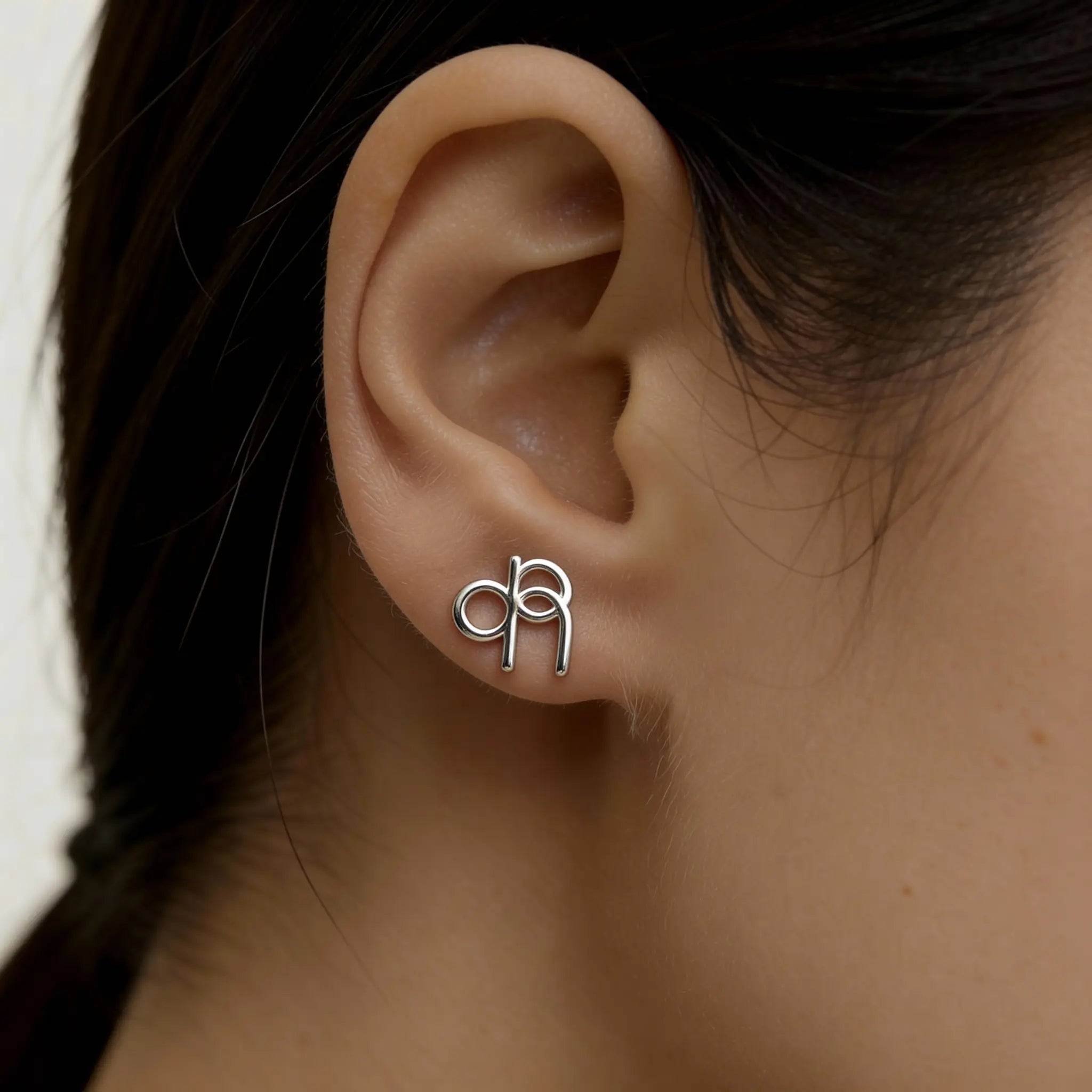Danibydsgn custom symbol stud earring in 925 sterling silver worn on ear by a lady,  featuring a cut-out silhouette of the design in lowercse letters.