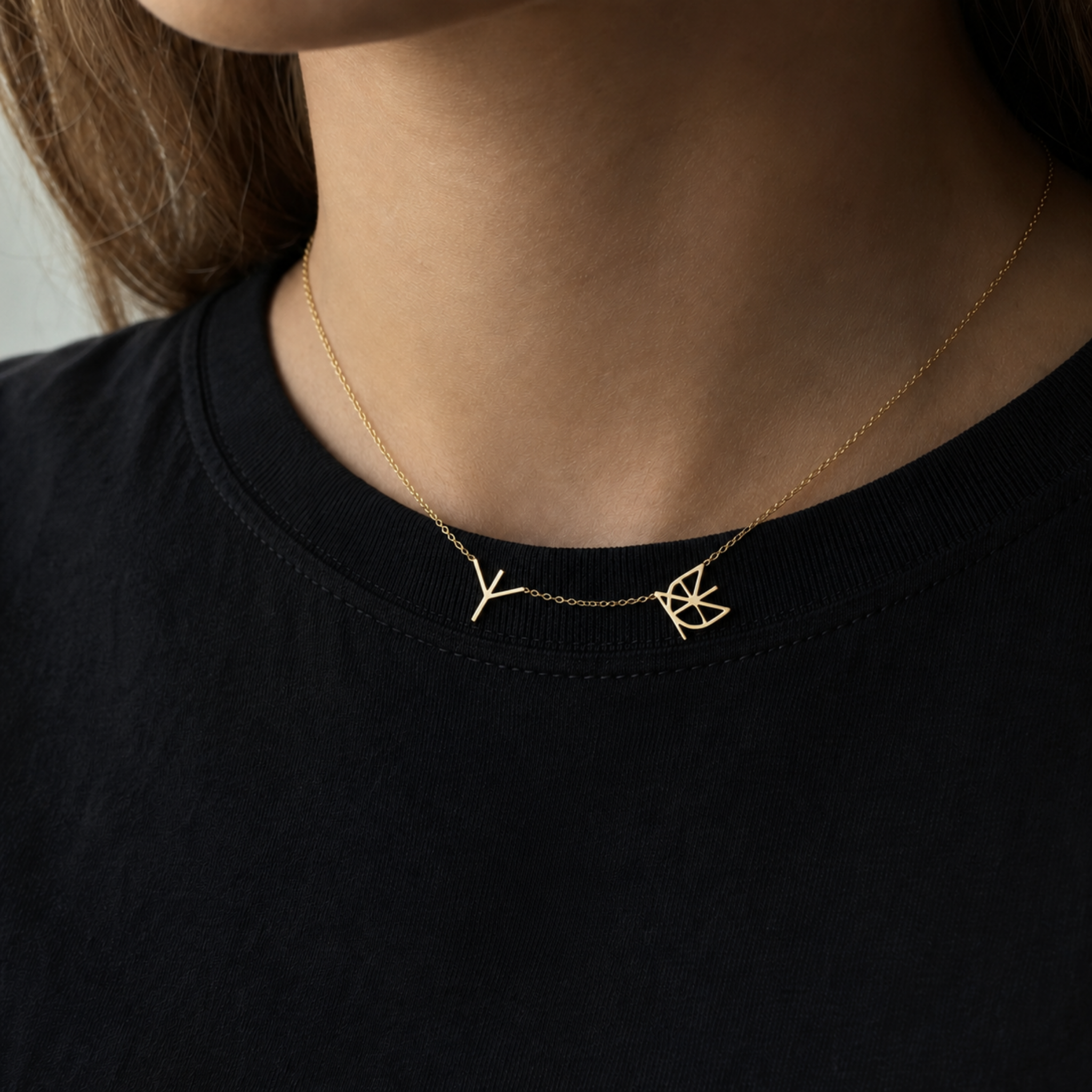 A close-up lifestyle shot of a model wearing a Danibydsgn two symbol pendant necklace in 9k solid gold. This made to order modern heirloom necklace features two personalized uppercase geometric charms handcrafted from a customer's unique names, words, or dates on a delicate yellow gold chain, styled against a black t-shirt.