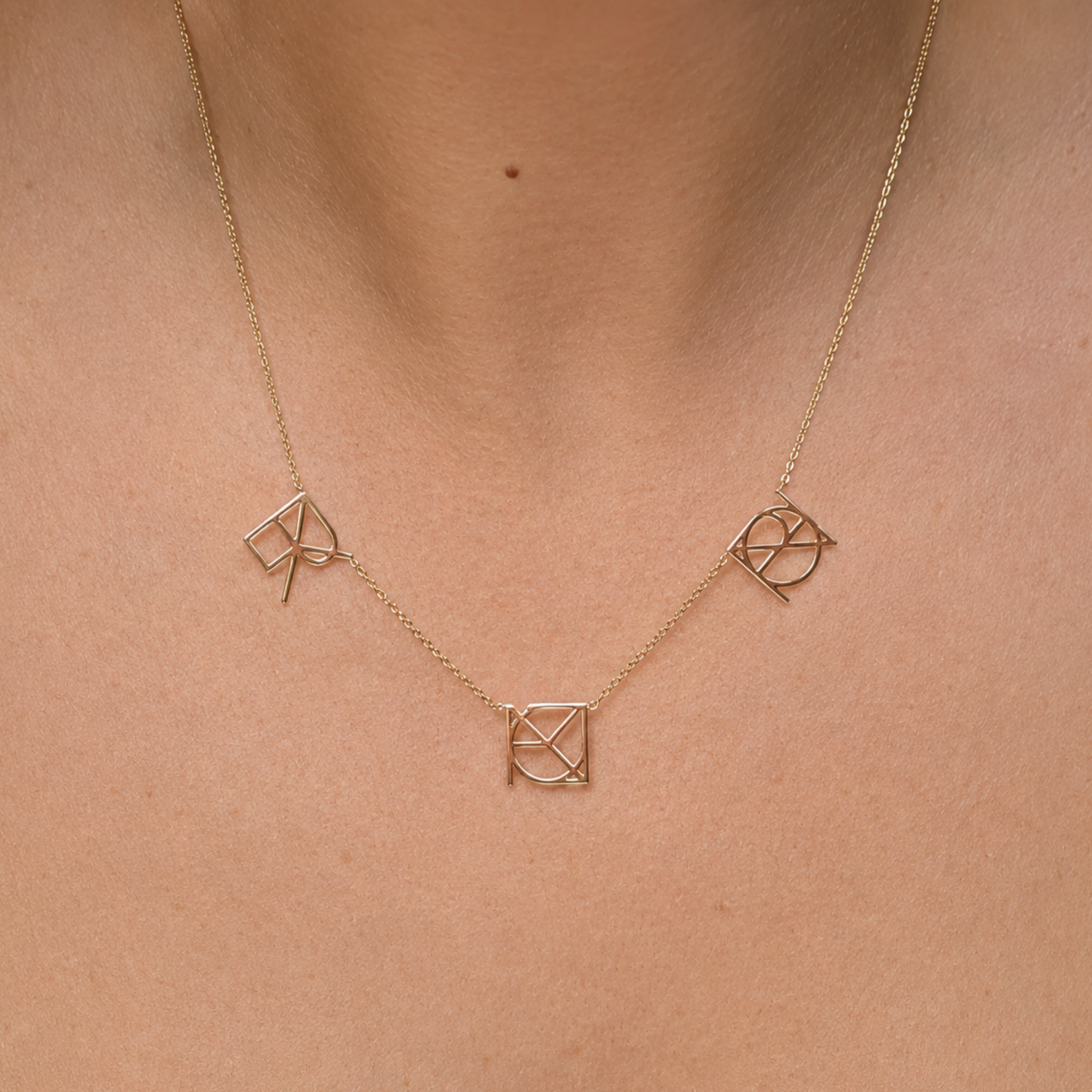 A close-up front view of a model wearing the Danibydsgn three symbol pendant necklace in 9k solid gold. This made to order modern heirloom necklace features three personalized uppercase geometric charms handcrafted from meaningful names, words, or dates, hanging beautifully on a delicate yellow gold chain against the skin.
