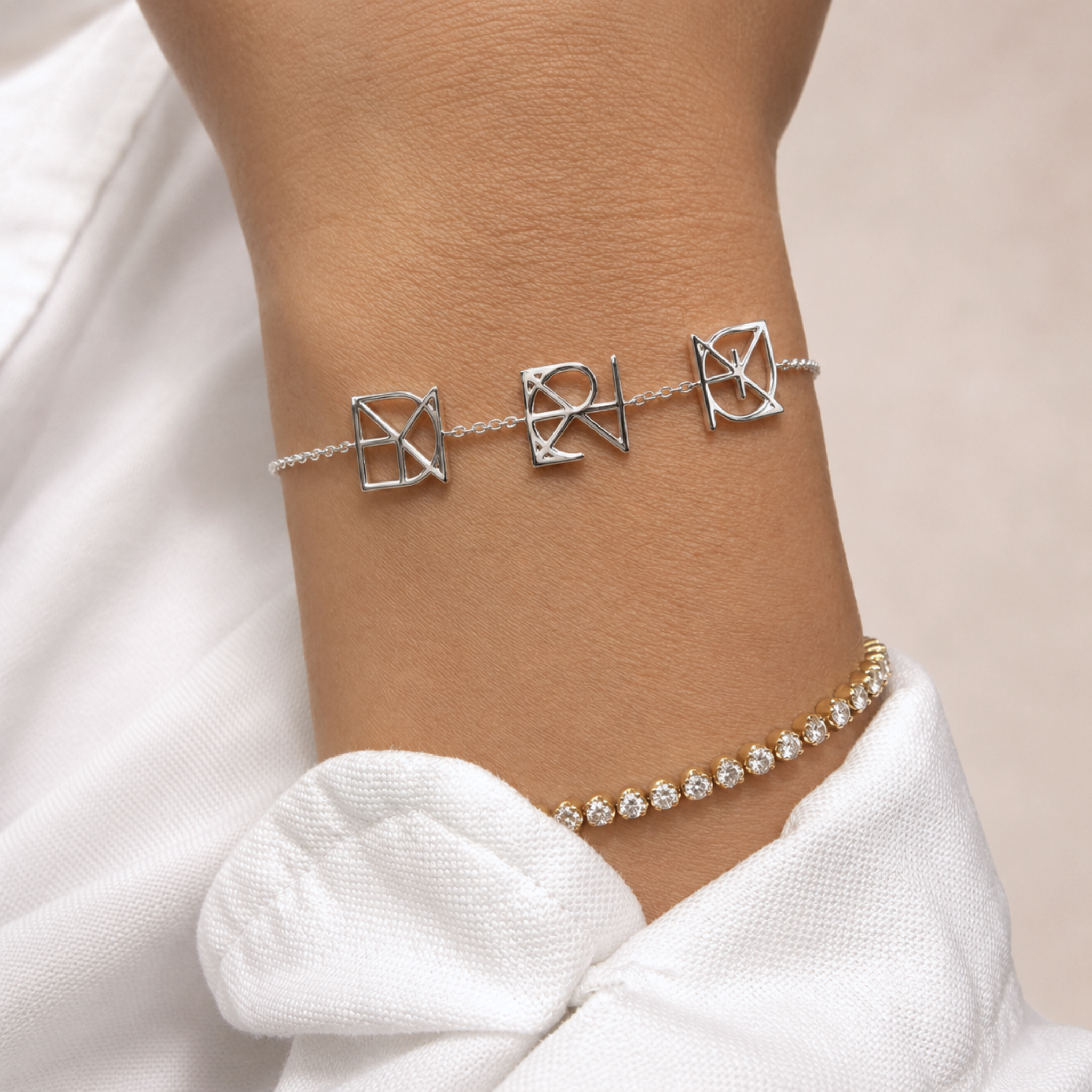 A close-up lifestyle shot of a model wearing a Danibydsgn three symbol pendant bracelet in 925 sterling silver. This custom line art charm bracelet features three uppercase Danibydsgn design, geometric square pendants on a delicate silver chain link wristlet, beautifully stacked on the wrist alongside a gold diamond tennis bracelet for a modern personalized jewelry look.