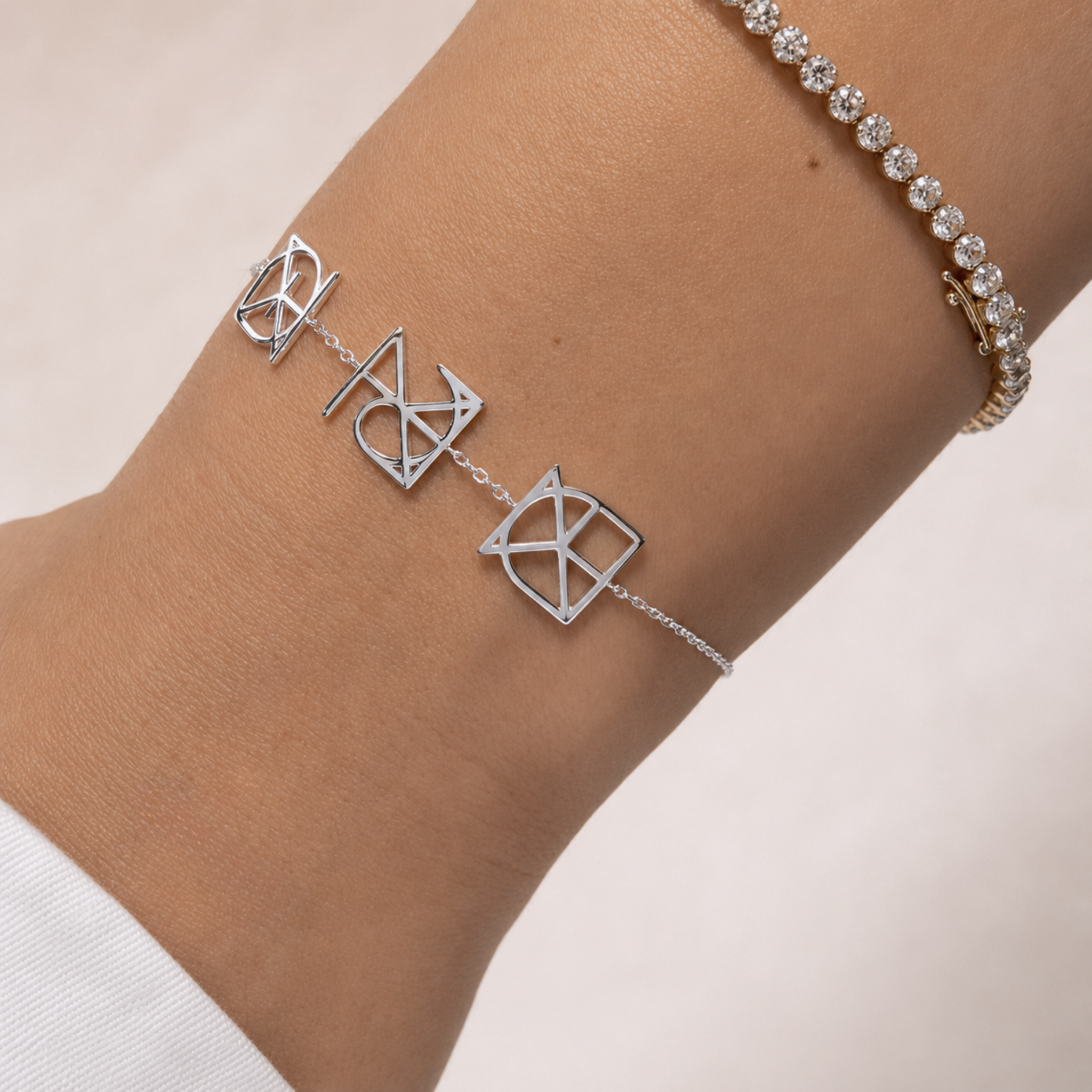 A close-up lifestyle shot of a model's wrist wearing a custom Danibydsgn three symbol pendant bracelet in 925 sterling silver. This made to order modern heirloom features three personalized uppercase geometric charms transformed from meaningful names, words, or dates, styled elegantly next to a gold diamond tennis bracelet.