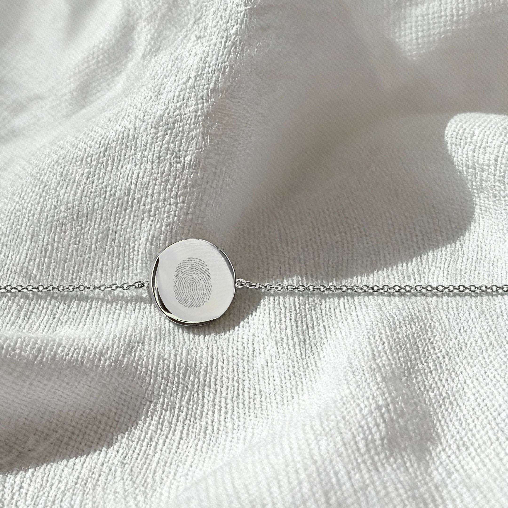 Elegant flatlay of the Danibydsgn Single Fingerprint Coin Bracelet in 925 Sterling Silver, displayed on softly textured white fabric, featuring a delicately engraved fingerprint on a polished circular pendant with a fine chain.