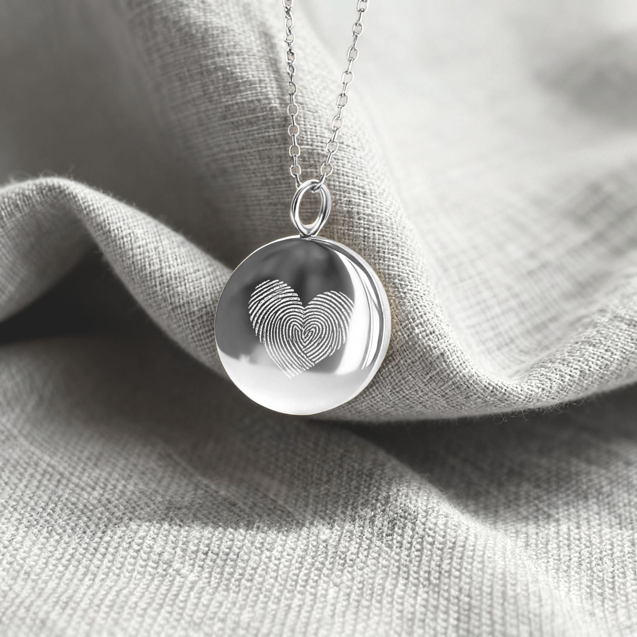 The Danibydsgn Heart Fingerprint Coin Necklace in 925 Sterling Silver resting on soft fabric — featuring two fingerprints engraved into a heart shape, symbolising love, unity, and timeless elegance in fine personalized jewelry.