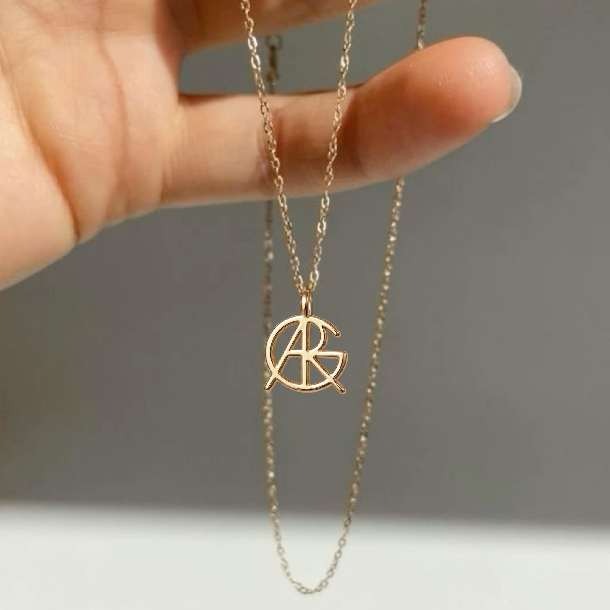 A close-up shot of fingers holding up a custom necklace in 9k gold, displaying a minimalist jewelry piece featuring a unique hand-designed Danibydsgn symbol cut out jewelry pendant with a top jump ring attachment.