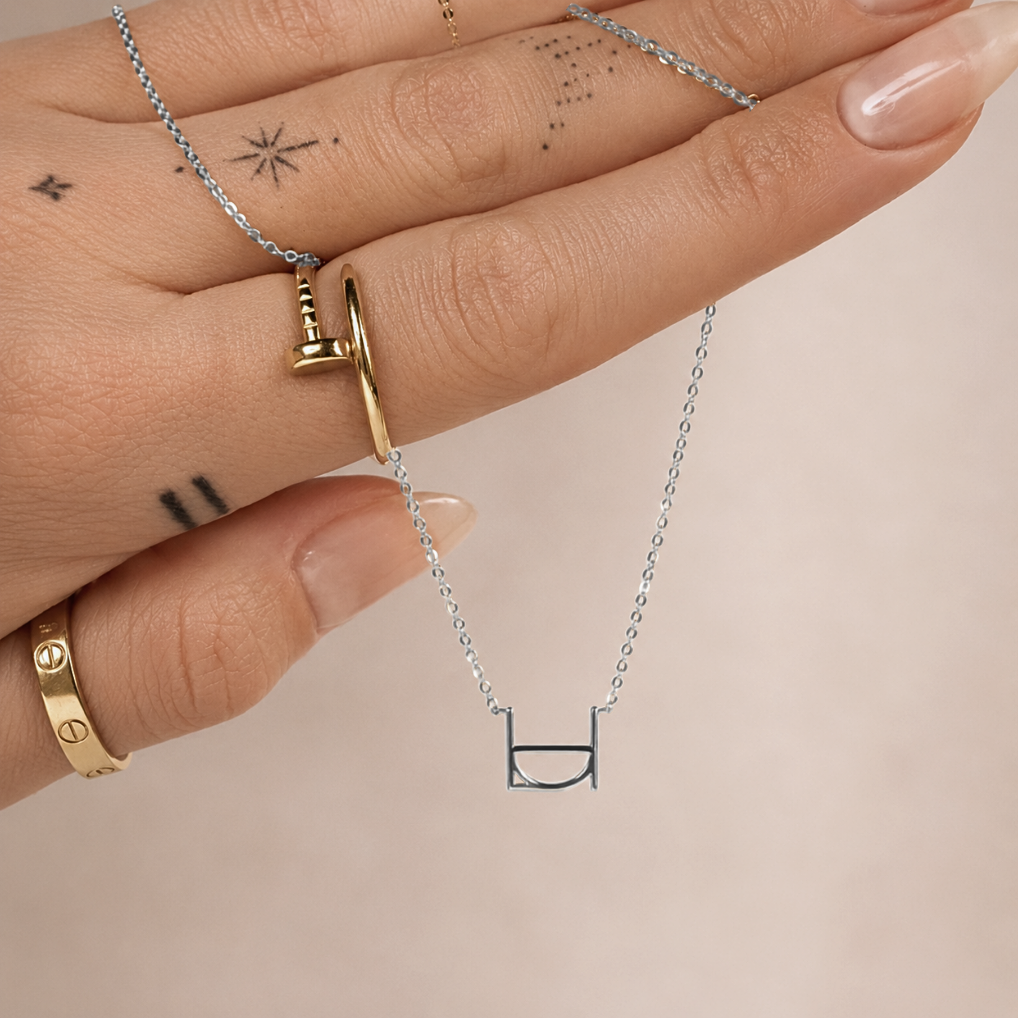 A close-up lifestyle shot of a model's hand displaying the Danibydsgn single symbol pendant necklace in 925 sterling silver. This dainty silver initial necklace features one personalized uppercase geometric charm handcrafted from a custom line art design, draped across tattooed fingers adorned with gold statement rings against a neutral background.