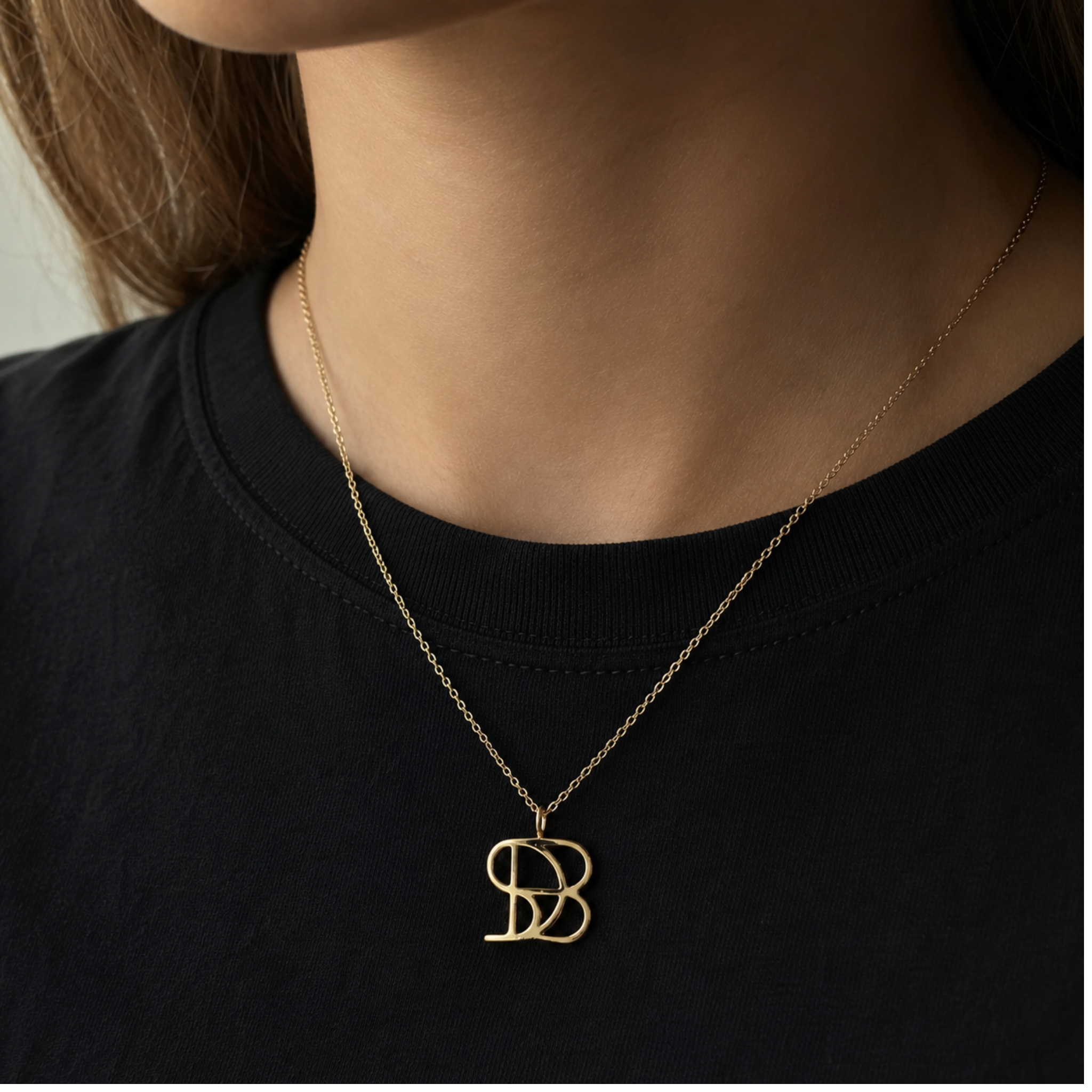 A close-up lifestyle image of a model wearing the Danibydsgn single symbol pendant necklace in 9k solid gold. This personalized architectural necklace features one custom uppercase geometric initial charm handcrafted from a fine-line design, elegantly resting on a delicate yellow gold chain against a classic black t-shirt.