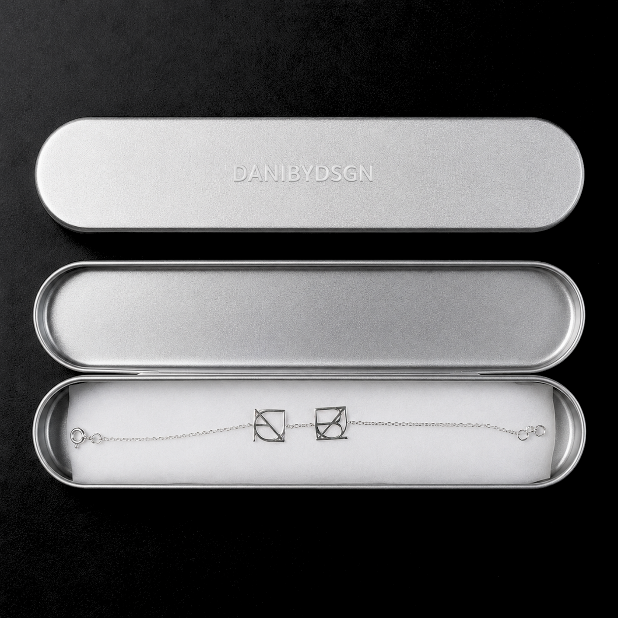 The Danibydsgn two symbol pendant bracelet in 925 sterling silver presented inside an open, brushed silver metal gift case. This personalized meaningful jewelry piece showcases two cut-out uppercase geometric charms handcrafted from a custom Danibydsgn design, resting flat on white padding next to the embossed brand name lid against a black background.