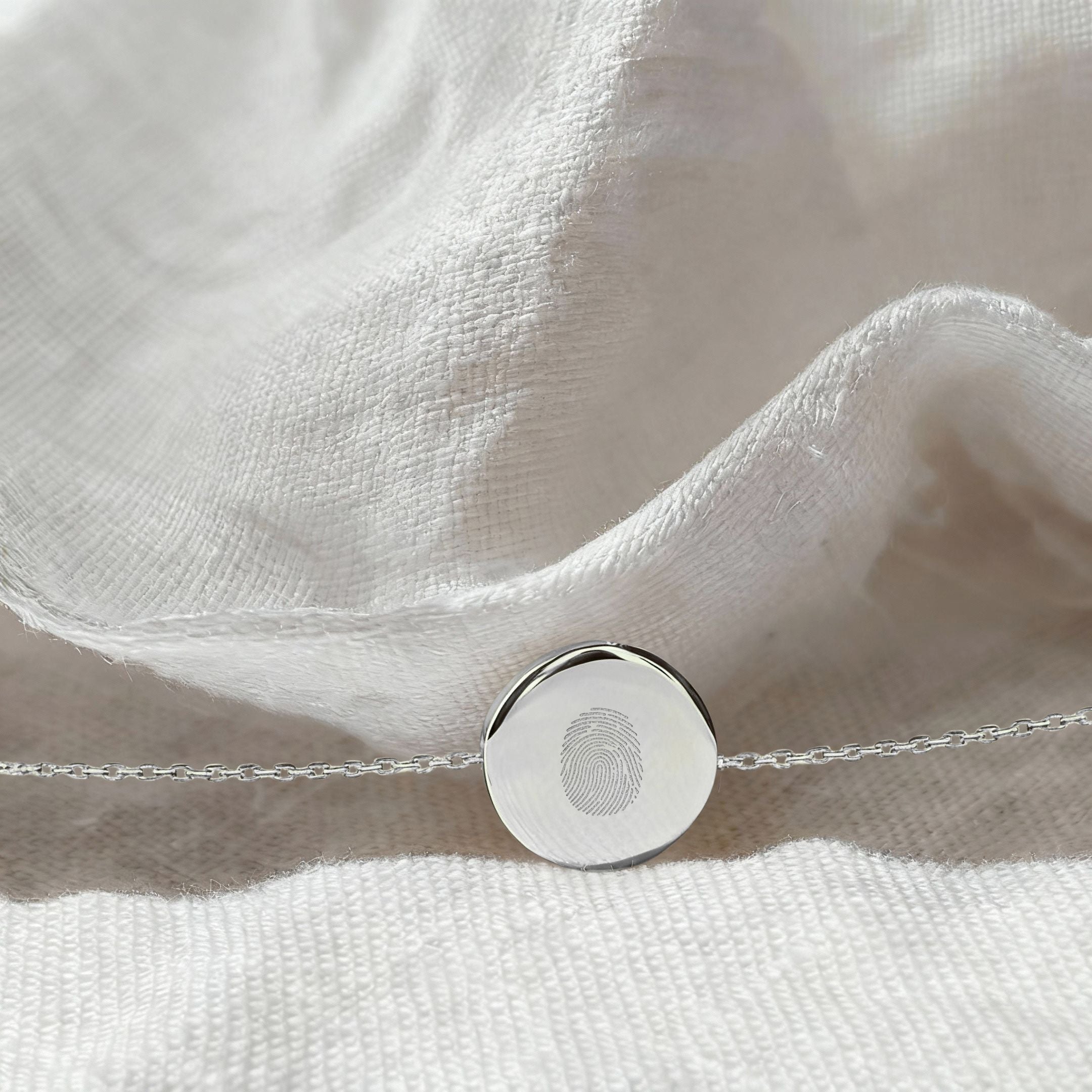 Elegant flatlay of the Danibydsgn Single Fingerprint Coin Bracelet in 925 Sterling Silver resting on white linen fabric, highlighting the engraved fingerprint pendant and delicate chain under soft natural light.