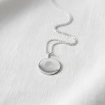 The Danibydsgn Single Fingerprint Coin Necklace in 925 Sterling Silver rests gracefully on a soft white fabric background. Its delicate chain and finely engraved fingerprint pendant reflect light with a serene, minimalist beauty — symbolising eternal love, memory, and connection in timeless silver craftsmanship.