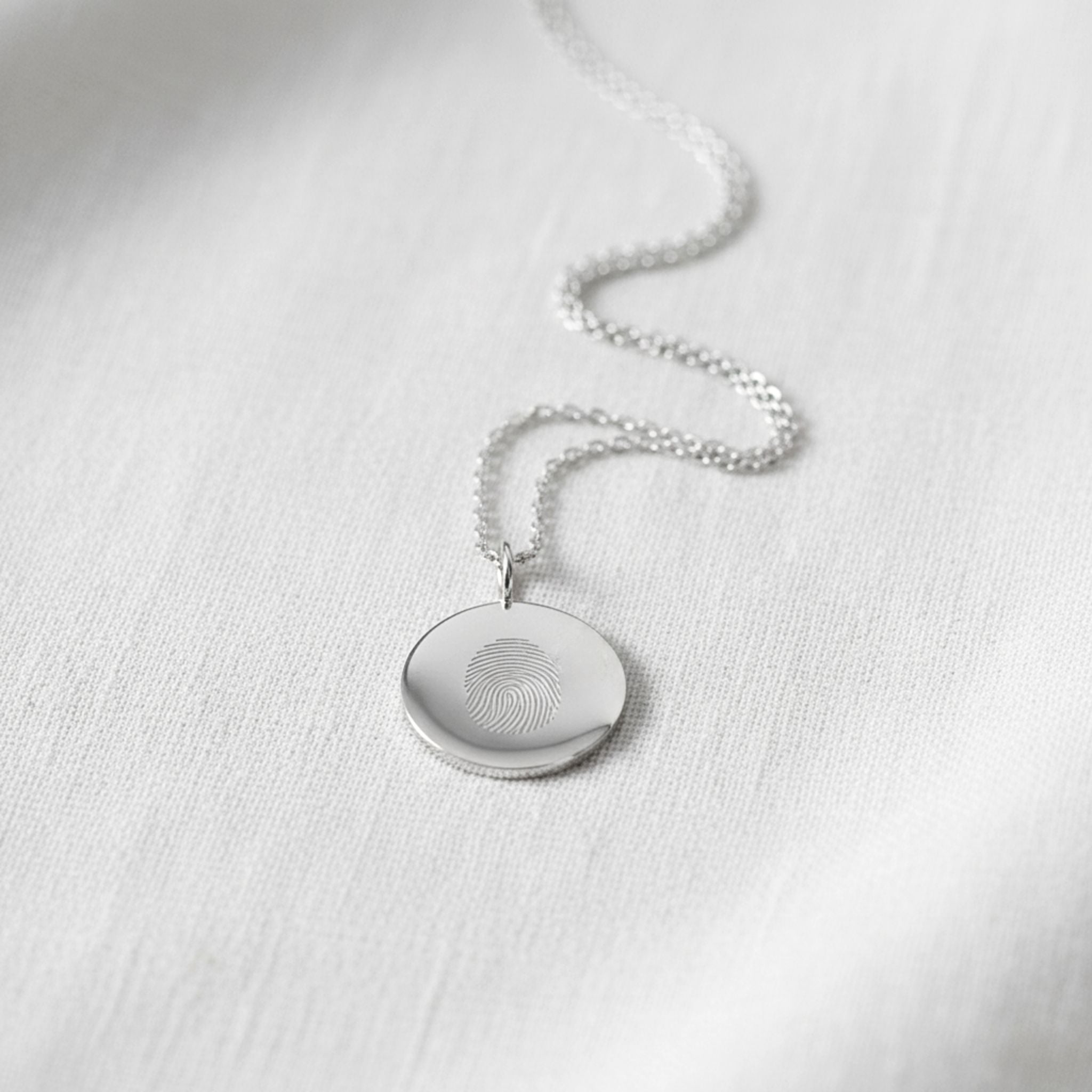 The Danibydsgn Single Fingerprint Coin Necklace in 925 Sterling Silver rests gracefully on a soft white fabric background. Its delicate chain and finely engraved fingerprint pendant reflect light with a serene, minimalist beauty — symbolising eternal love, memory, and connection in timeless silver craftsmanship.