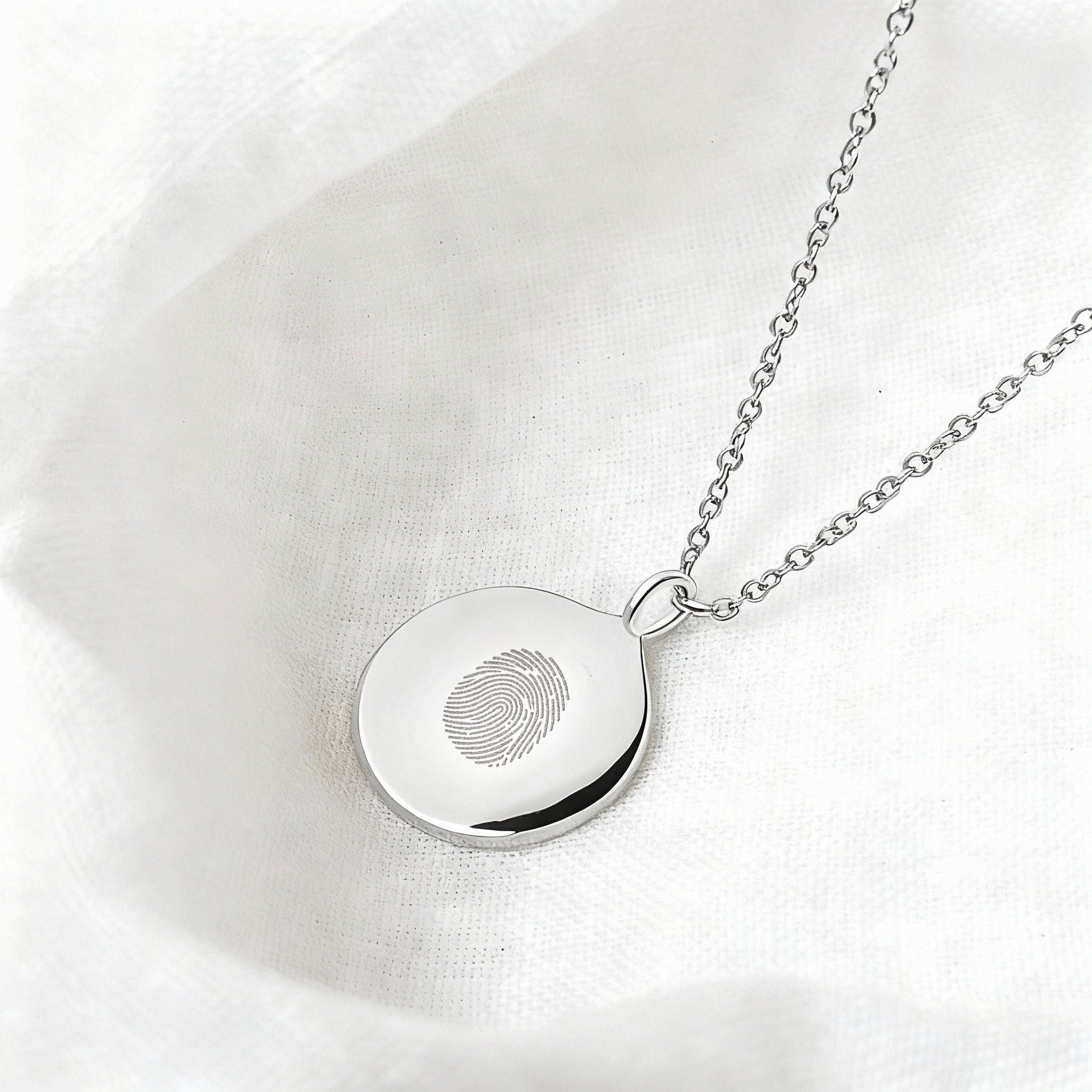 The Danibydsgn Single Fingerprint Coin Necklace in 925 Sterling Silver, resting elegantly on soft white fabric. Its polished surface reflects light beautifully, highlighting the engraved fingerprint — a delicate expression of love, remembrance, and connection.
