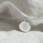 The Danibydsgn Single Fingerprint Coin Necklace in 925 Sterling Silver, elegantly displayed on textured white fabric. Its sleek, mirror-like finish and engraved fingerprint create a modern yet sentimental keepsake — a lasting tribute to love and memory.