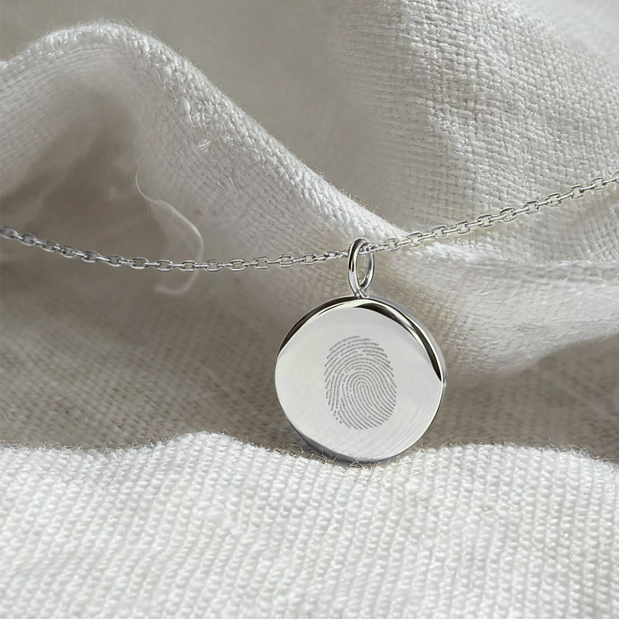 The Danibydsgn Single Fingerprint Coin Necklace in 925 Sterling Silver, elegantly displayed on textured white fabric. Its sleek, mirror-like finish and engraved fingerprint create a modern yet sentimental keepsake — a lasting tribute to love and memory.