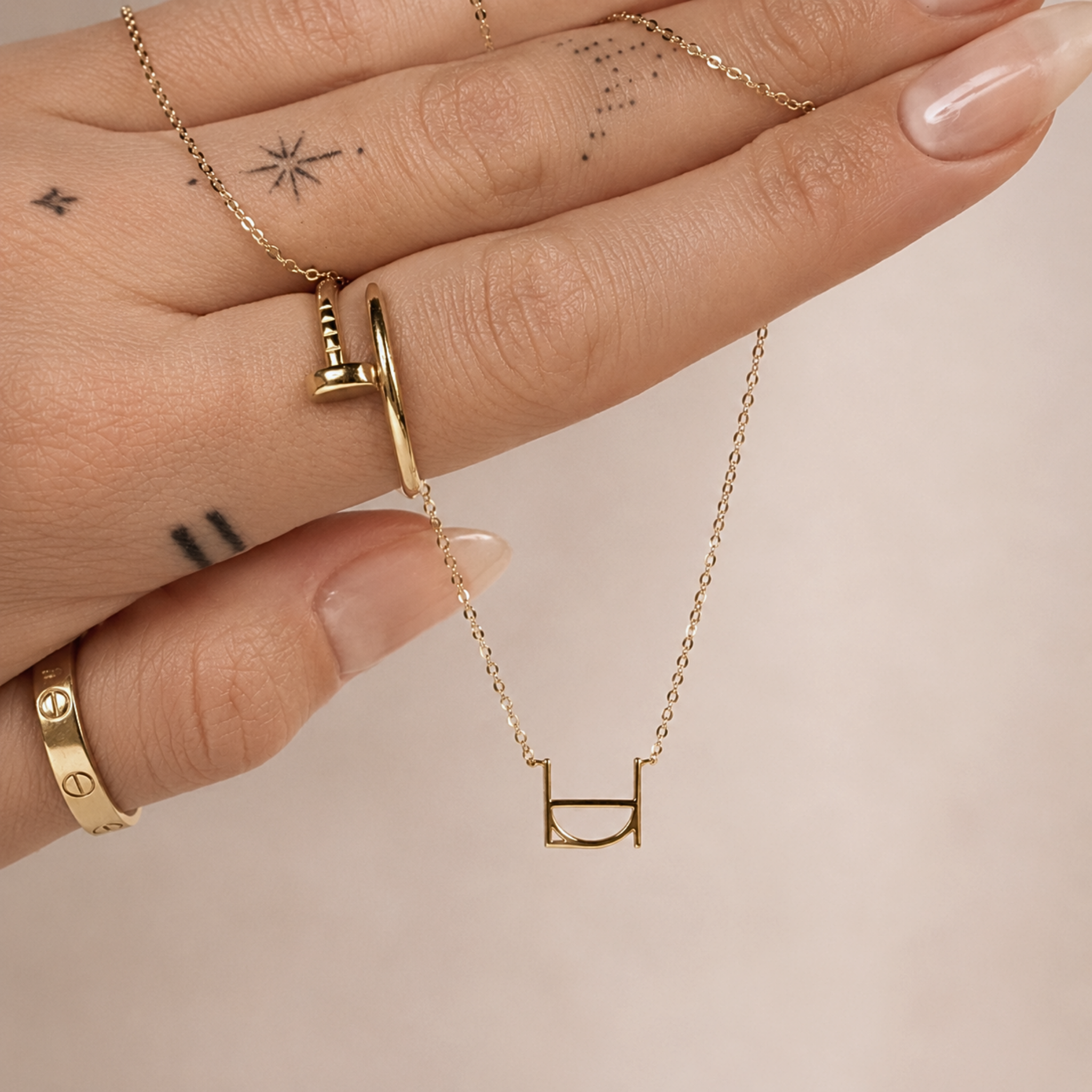 A close-up lifestyle shot of a model's hand displaying the Danibydsgn single symbol pendant necklace in 9k solid gold. This dainty gold initial necklace features one personalized uppercase geometric charm handcrafted from a custom line art design, draped elegantly across tattooed fingers adorned with gold statement rings against a neutral background.