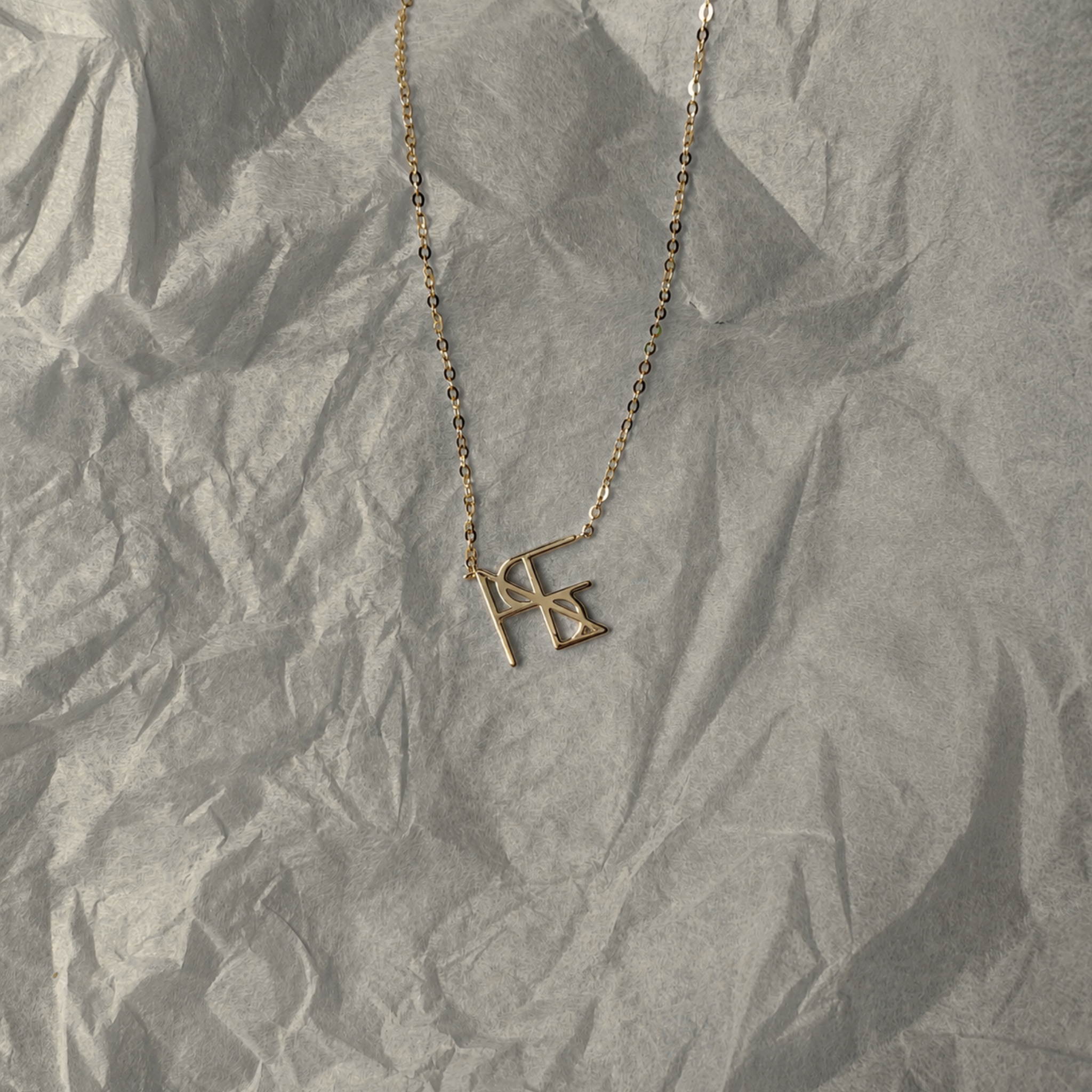 A top-down studio product flat-lay of the Danibydsgn single symbol pendant necklace in 9k solid gold. This personalized fine jewelry piece features a custom uppercase geometric monogram charm on a delicate yellow gold chain, laid out beautifully on a textured background of crinkled white tissue paper.