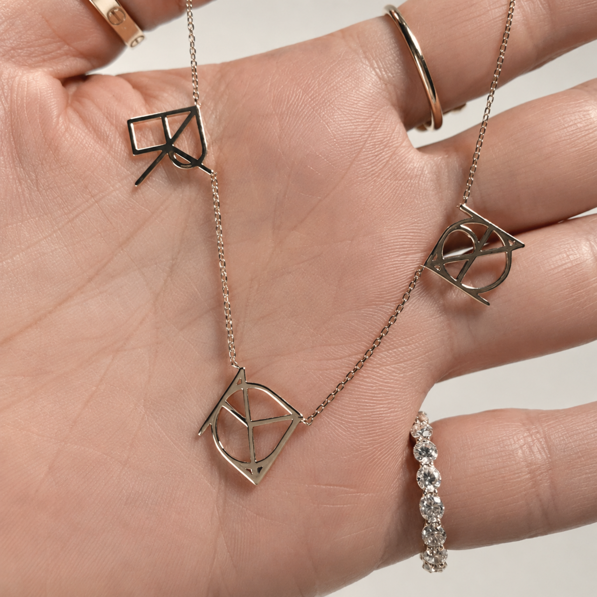 A detailed close-up shot of a model's open palm holding the Danibydsgn three symbol pendant necklace in 9k solid gold. This made to order modern heirloom highlights three personalized uppercase geometric charms handcrafted from meaningful names, words, or dates, draped across the hand to display the fine-line cut-out details of the yellow gold charms.